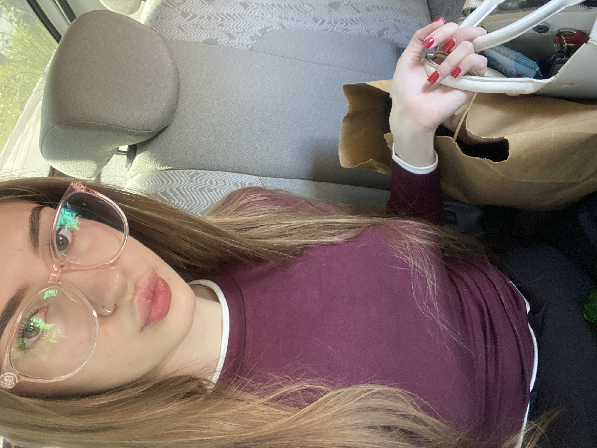 This media file captures a selfie of a young woman, taken from a high angle and presented rotated 90 degrees clockwise. The woman, occupying the left side of the frame, has long, light brown hair that cascades over her shoulders. She wears large, light pink-framed glasses, whose lenses reflect a vibrant green and rainbow-like shimmer. A small silver hoop pierces her left nostril, and her lips are slightly parted in a gentle pout, appearing to be enhanced with lip gloss or lipstick. She is dressed in a maroon long-sleeved shirt with a distinct white trim around the collar.
Her right hand, featuring bright red nail polish, grasps the white handle of a purse. Inside the open purse, a blue packet and what appear to be keys are visible. Adjacent to the purse, a brown paper bag is partially shown.
The background clearly depicts the interior of a car, with a gray fabric car seat and headrest visible. The seat fabric has a subtle, light gray geometric pattern. Bright ambient light, likely sunlight, enters from the upper left, suggesting it is daytime. The image portrays a casual moment, possibly while traveling or waiting in a vehicle. There is no discernible text within the image.