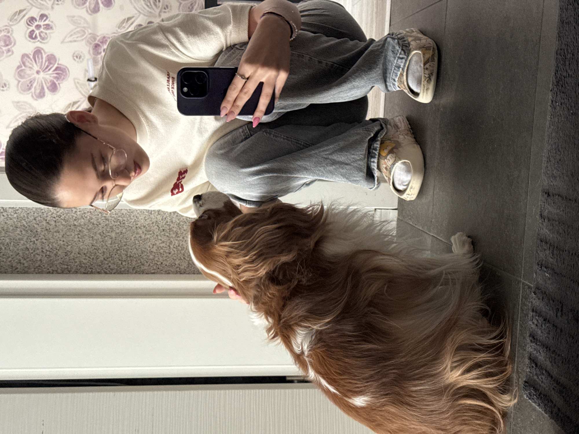 The image shows a young woman wearing glasses and a light-colored sweatshirt, kneeling on a tiled floor indoors. She is holding a dark smartphone in her right hand and gently petting a brown and white dog with her left hand. The dog is a Cavalier King Charles Spaniel, facing the woman. The background features a white door, a textured wall with a floral pattern, and a dark rug in the foreground. There is no indication of the location being Sofia, Bulgaria.