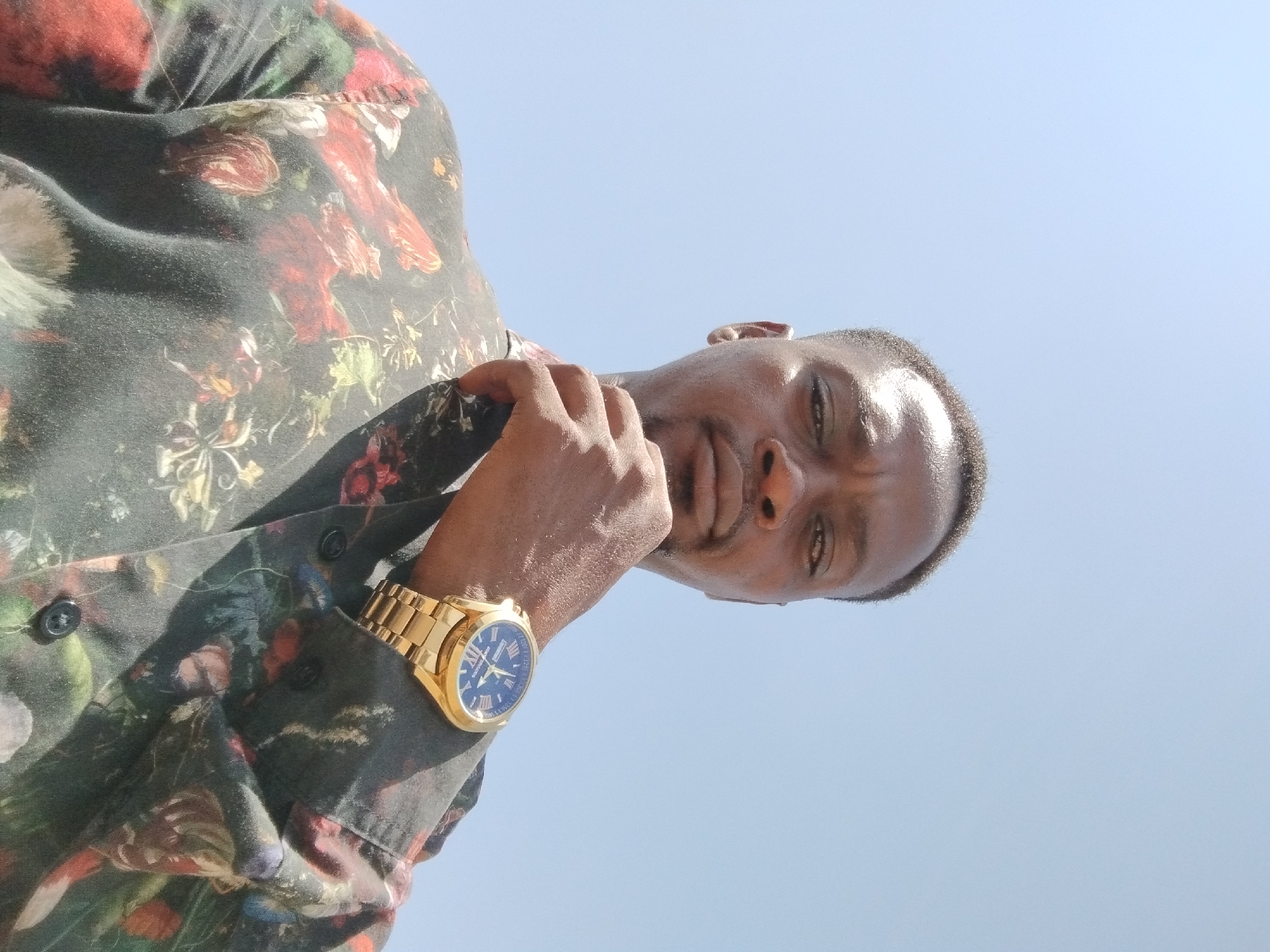 A man is pictured from a low angle against a clear blue sky. He is wearing a button-down shirt with a dark background and a busy floral pattern in shades of red, green, and cream. A gold wristwatch with a blue face is visible on his left wrist. His left hand is raised towards his face, with his fingers near his chin. The location is New Stadium, Nigeria.
