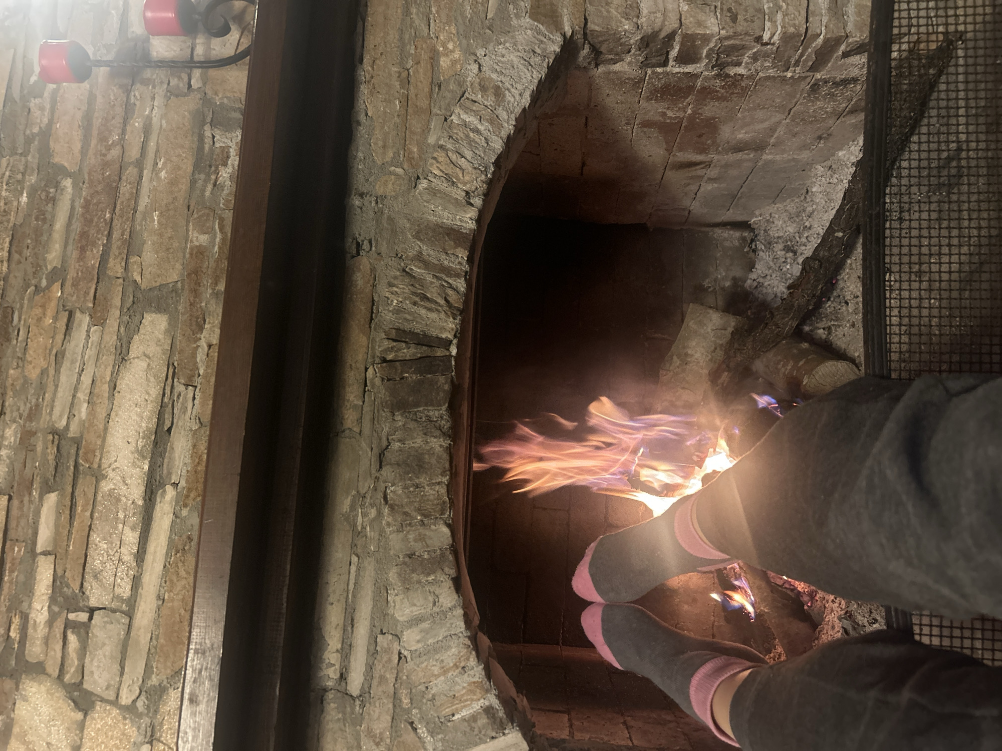 The image shows a stone fireplace with a fire burning inside. A person wearing grey socks with pink stripes is sitting in front of the fireplace. The walls surrounding the fireplace are made of irregular stone bricks. A metal grate is partially visible on the right side of the fireplace interior. There is no context in the image to determine the location of Dolen, Bulgaria.