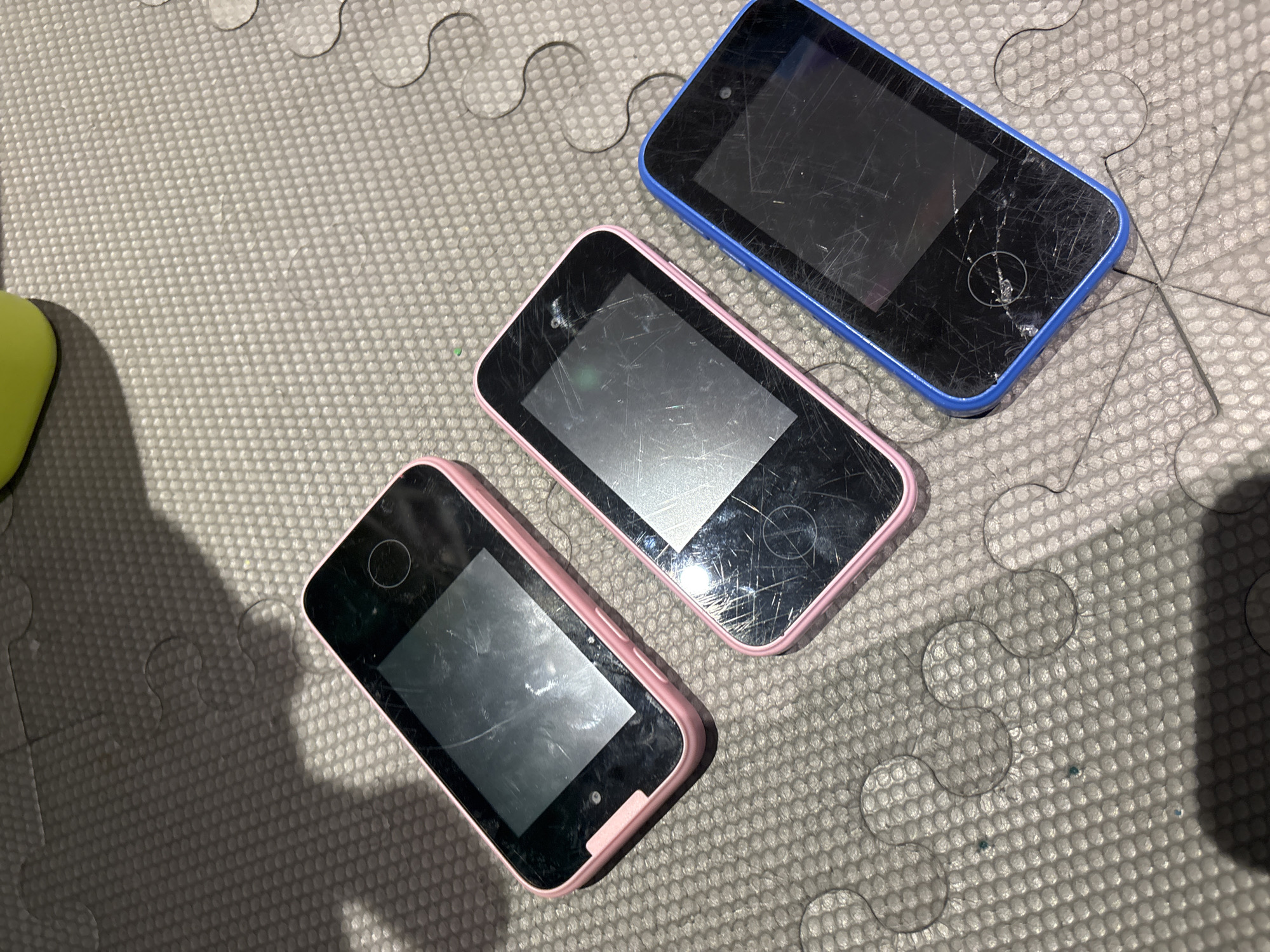 Three scratched mobile phones are placed on a grey, textured mat. The phones are arranged diagonally from the top left to the bottom right of the image. The phone on the far left is pink, the middle phone is also pink, and the phone on the far right is blue. The mat has a pattern of small raised circles and interlocking shapes, resembling a puzzle mat. A faint shadow is cast across the mat from the upper left. The scene is indoors, on a mat likely used for play or exercise. There are no people or specific identifying landmarks of Dubai, United Arab Emirates visible in the image.