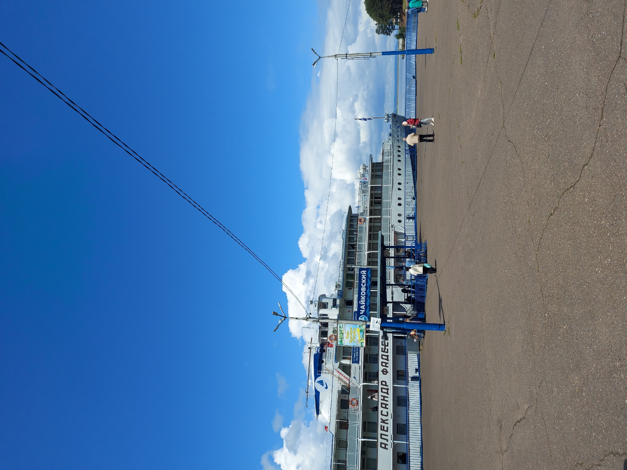 The image depicts the cruise ship "Aleksandr Fadeev" docked at the pier in the city of Chaikovsky, Russia. The ship is white with blue accents and its name is written in Cyrillic on the side. A sign with the name "Chaikovsky" is also visible. Several people are present near the ship and on the pier. The sky is blue with scattered clouds. The pier is made of asphalt. Power lines are seen stretching across the sky.