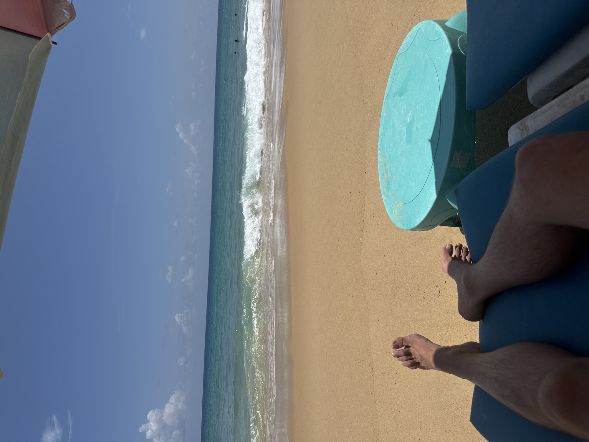 A first-person perspective shot shows a person's legs and feet resting on a blue lounge chair on a sandy beach. A small, round, teal-colored table sits to the right of the chair. The ocean is visible in the background with waves breaking on the shore under a clear blue sky with scattered clouds. The location is Karon Beach in Karon, Thailand.
