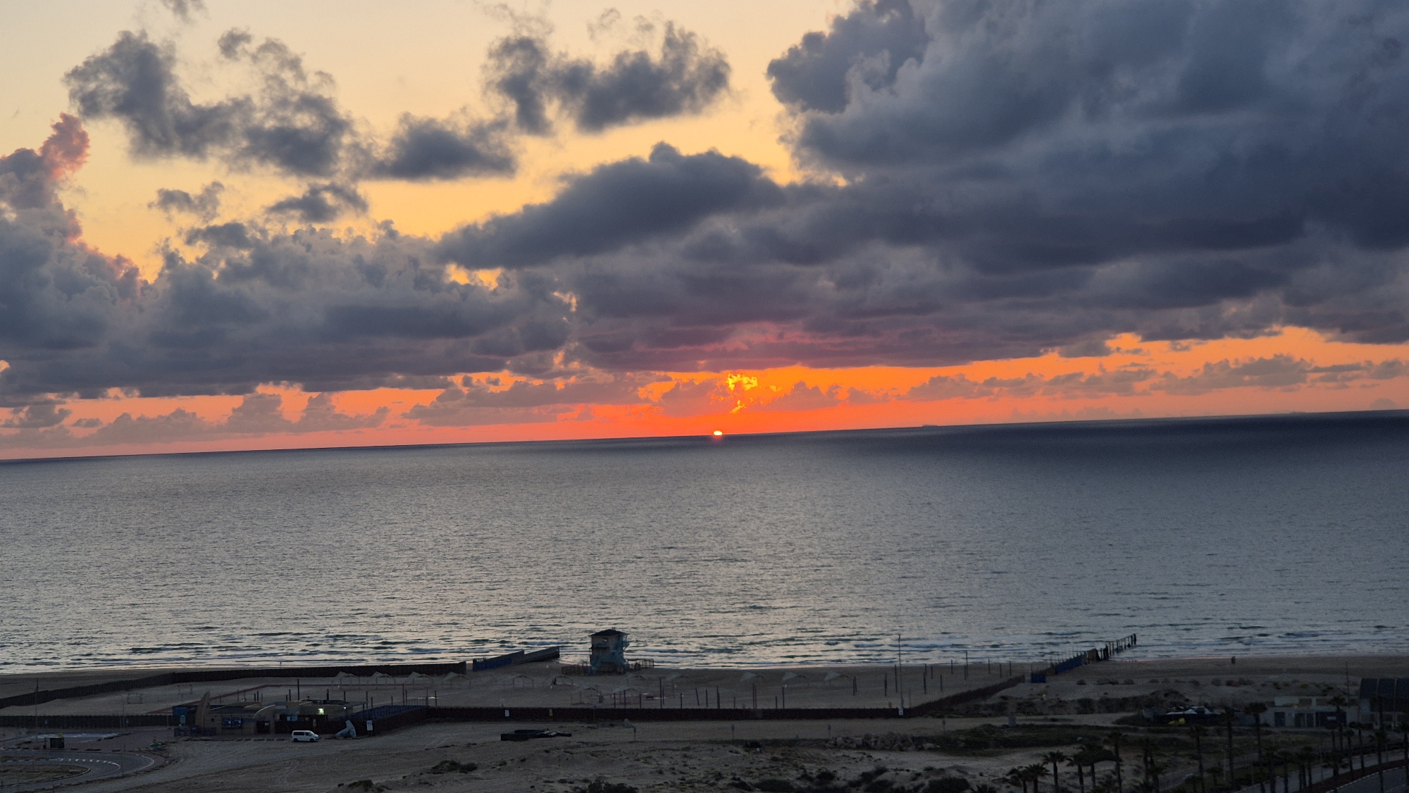The image captures a sunset over the sea in an unknown city and country. The sky is filled with dark, gray clouds at the top, with some tinged with orange and pink from the setting sun. A narrow horizontal band of intense orange light stretches across the horizon where the sun is partially obscured by the clouds, creating a bright glow. Below the horizon, the sea is calm with gentle waves reflecting the light from the sky. The beach area is visible in the foreground with some structures and buildings, including a lifeguard station, and some vehicles parked nearby. A sandy area leads to the beach, with palm trees and other vegetation visible at the edge of the frame.