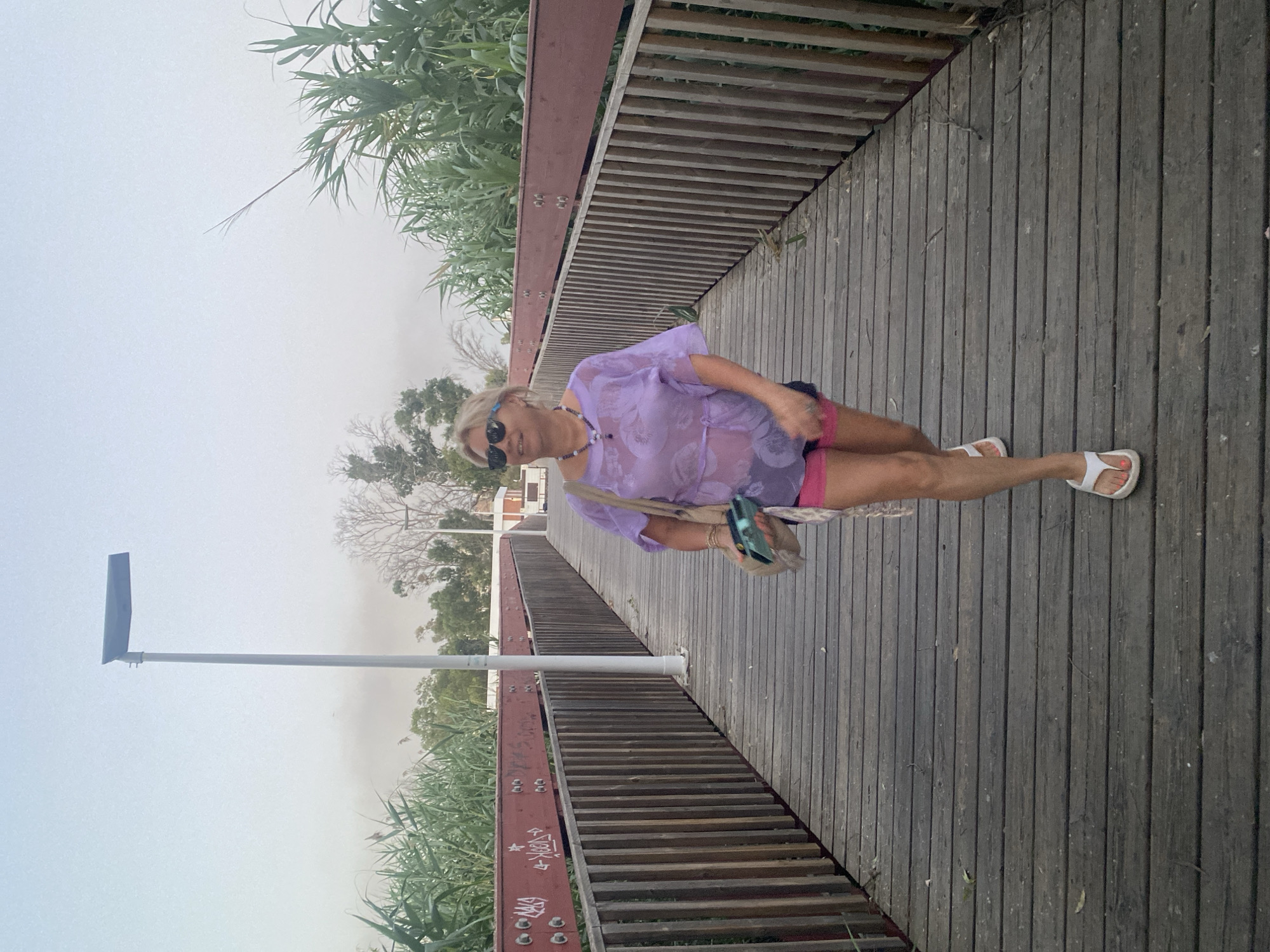 A woman walks on a wooden boardwalk lined with wooden railings and lush greenery. She wears a sheer purple blouse, pink shorts, and white flip-flops. The boardwalk appears to be in a park or natural area, with a lamppost visible in the background. The sky is overcast, suggesting a cloudy day. The location is identified as Resinera-voladilla, Spain.