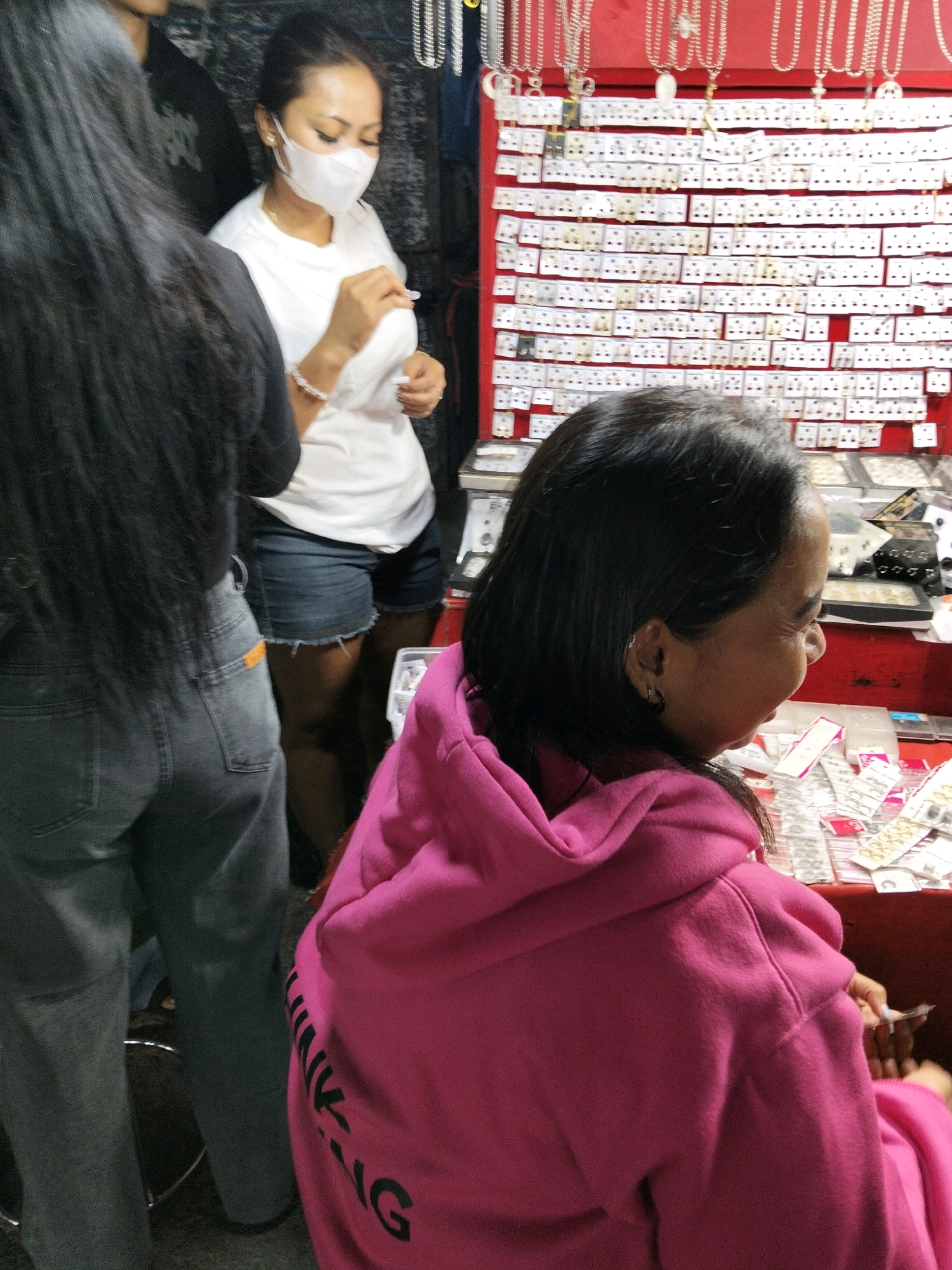 In Denpasar, Indonesia, at a street market stall, a woman wearing a pink hooded sweatshirt is looking at a display of jewelry. The stall features numerous small necklaces and earrings mounted on white cards. To her left, another woman in a white t-shirt and denim shorts stands, wearing a white face mask and examining a piece of jewelry. Her left arm is adorned with a bracelet. Behind her, a person with long black hair is partially visible, wearing a dark shirt and grey jeans. The market stall itself is brightly lit by overhead lights, illuminating the rows of merchandise and the faces of the shoppers. The background is dark and somewhat obscured, typical of an outdoor market setting.