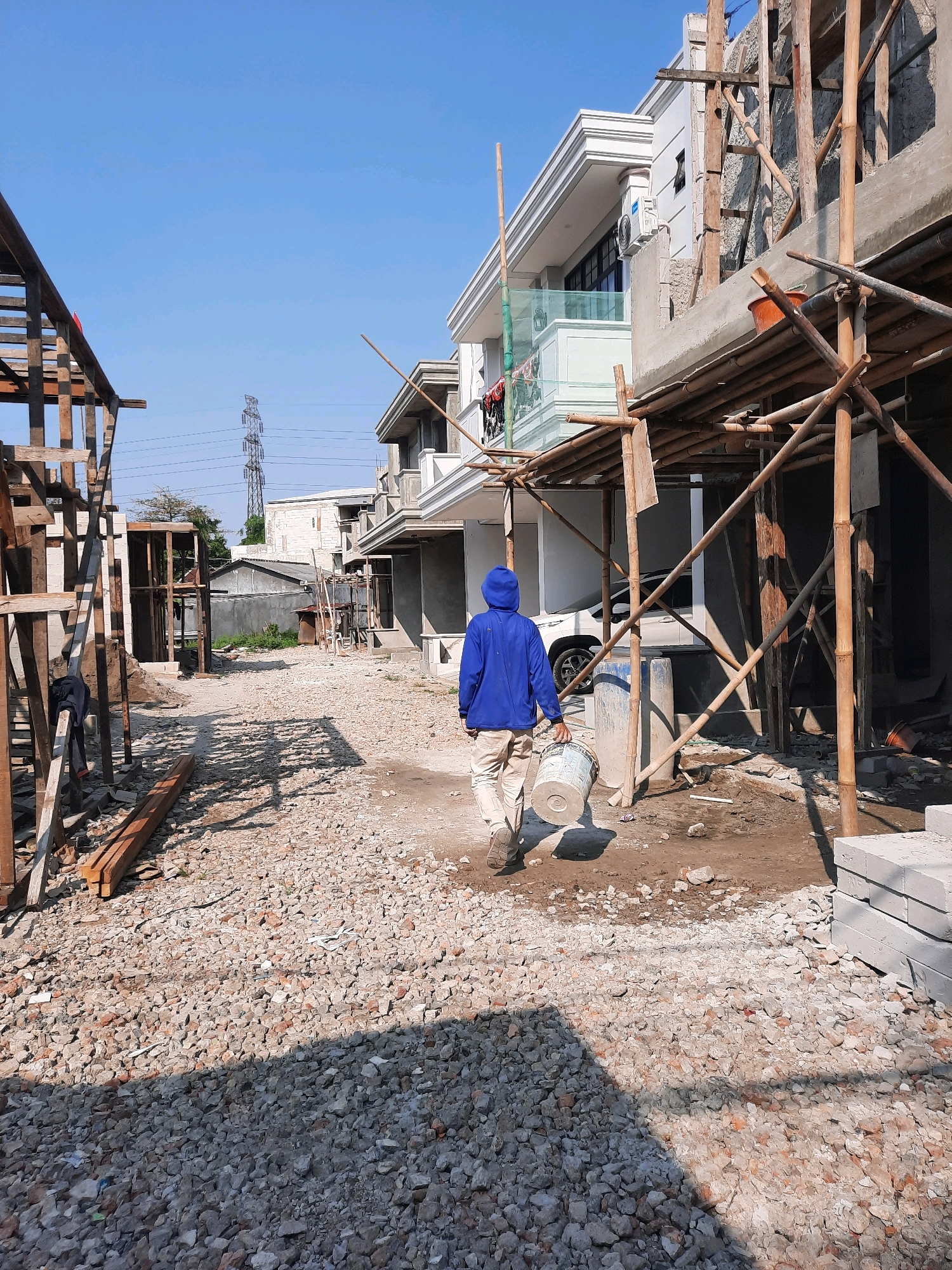 A male construction worker wearing a blue hoodie and light-colored pants walks away from the camera carrying a white bucket. He is on a path covered in gravel and debris. To the left of the frame is a partially constructed wooden structure, and to the right is a modern house under construction, with scaffolding made of bamboo. In the background, there are other houses and a tall transmission tower against a clear blue sky. The scene appears to be in a developing residential area in East Jakarta, Indonesia, characterized by ongoing construction activities.