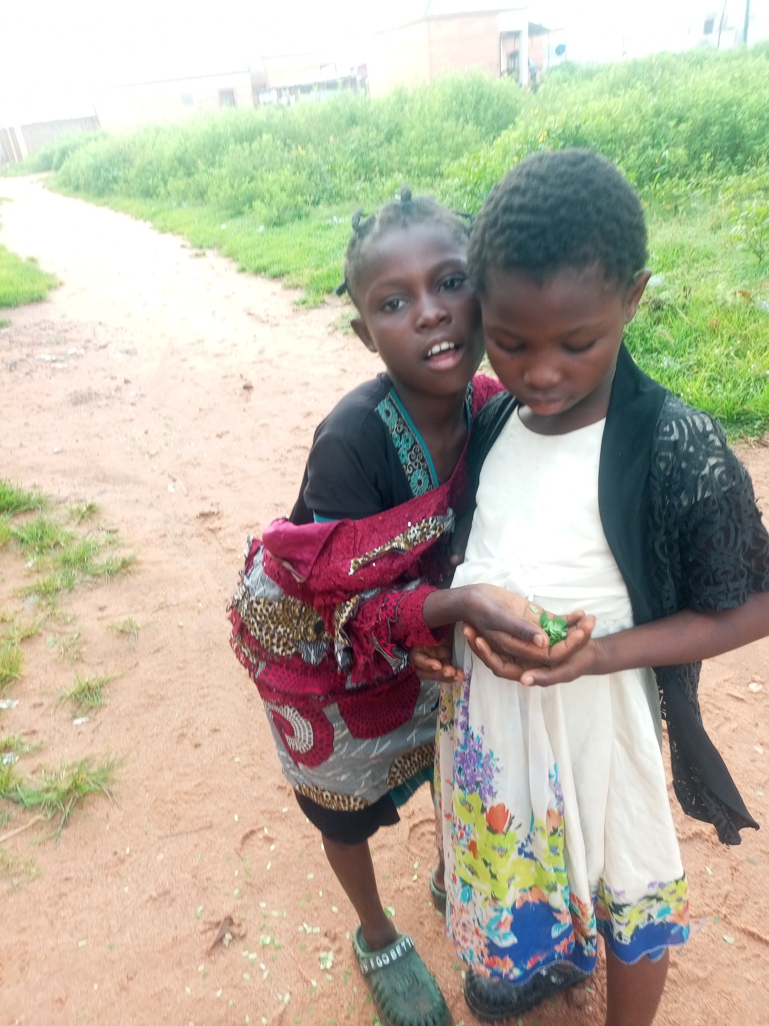 Two young girls stand close together outdoors on a dirt path. The girl on the left, with her hair in two braids, wears a dark top and a patterned burgundy and leopard-print skirt. She is looking directly at the camera with her mouth slightly open. The girl on the right, with short dark hair, is looking down at her hands, which hold a small cluster of green leaves. She wears a white dress with a floral print and a black lace overlay on her shoulders. The background shows a grassy embankment and a hazy, light-colored sky, suggesting a cloudy day. The scene appears to be in Tempure, Nigeria.