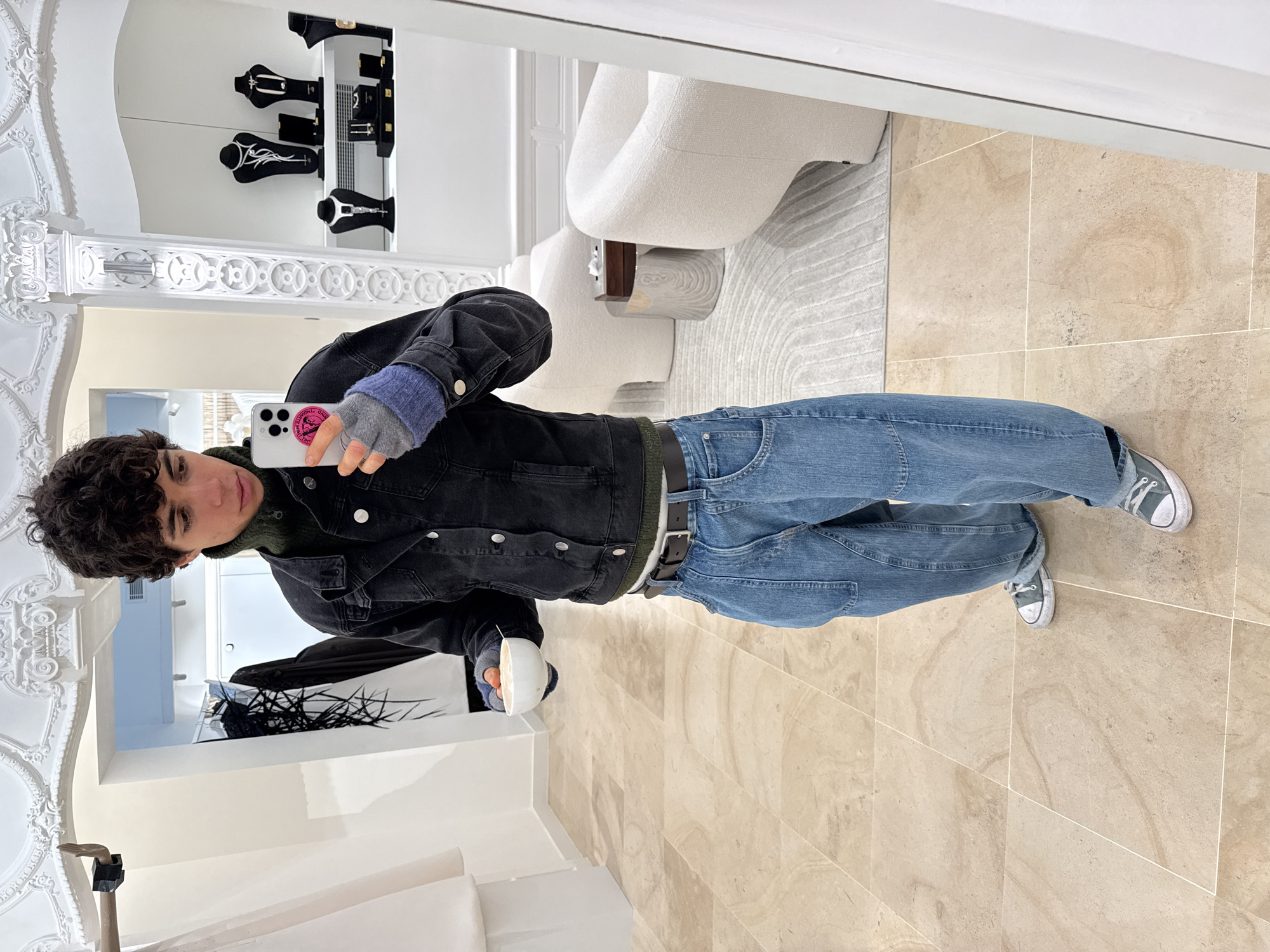 A person stands in what appears to be a high-end retail space. They are taking a selfie in a mirror. The individual is wearing a black denim jacket over a dark green sweater, loose light blue jeans, and gray sneakers. They have curly dark hair and are holding a white smartphone in their left hand.
Behind them, the retail space features a light-colored floor, modern white furniture, and display cases showcasing jewelry. The location has ornate, classical architectural details, including a decorative ceiling molding. Based on the upscale appearance, it's plausible that the image was taken in a luxury retail district, though the exact street or shop cannot be determined from the image alone.