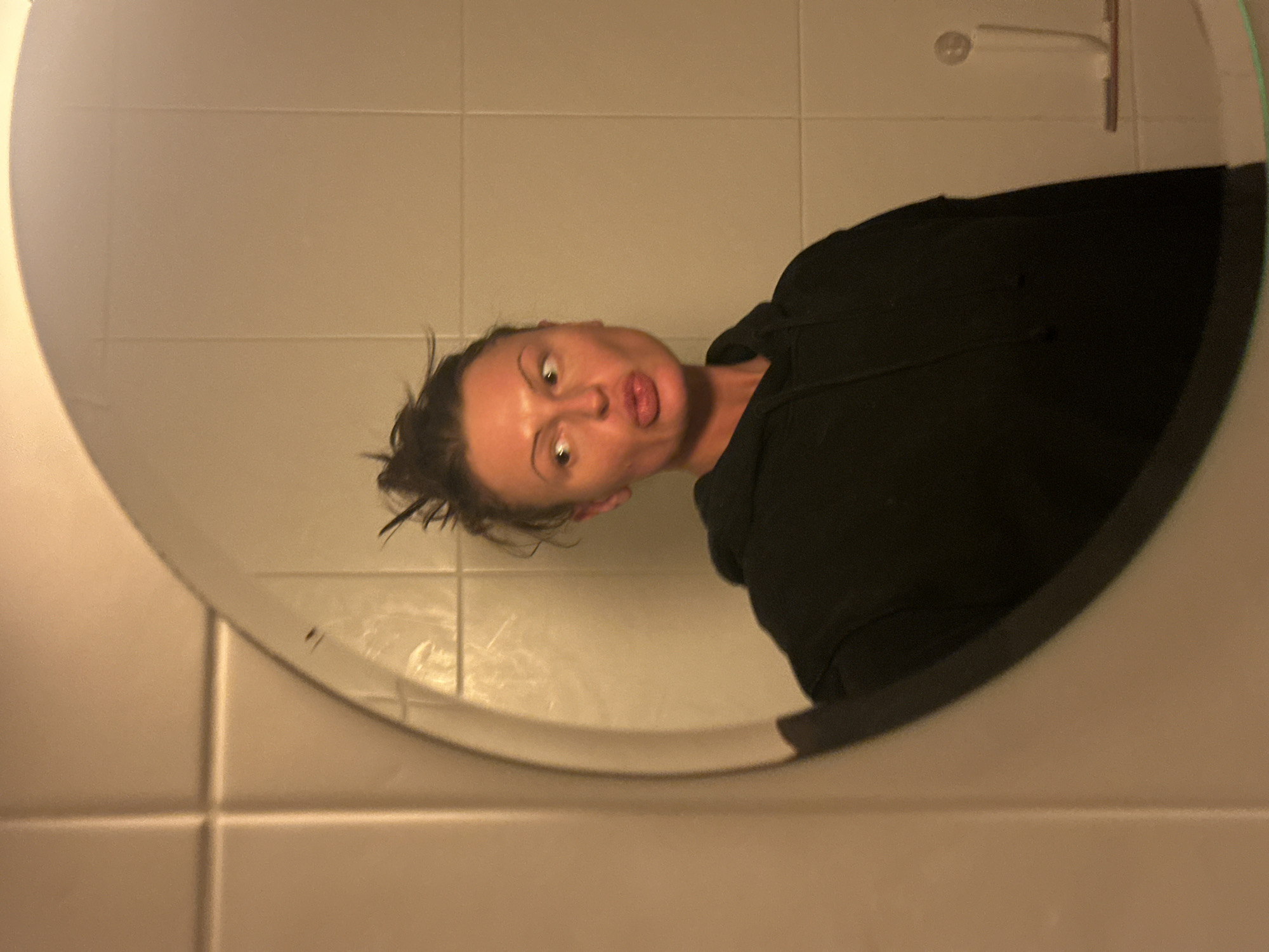 A woman with her hair up and wearing a black hoodie is reflected in a circular mirror. The background is a wall covered in beige tiles. A squeegee is visible above the mirror. The location appears to be a bathroom. The image does not contain enough information to confirm the location within Tunbridge Wells, United Kingdom.