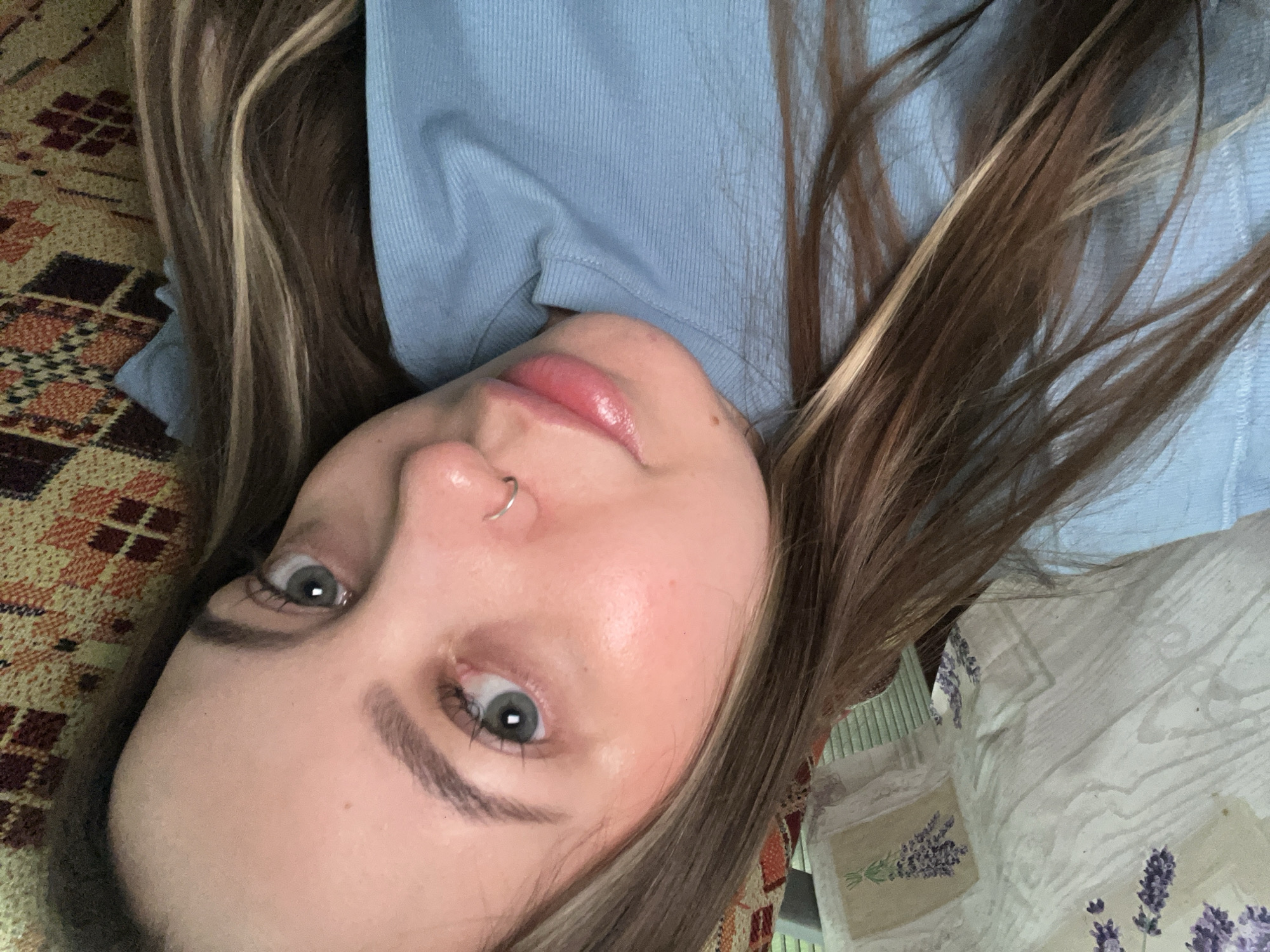 A young woman with brown hair and blue eyes is captured in a close-up selfie. She is wearing a light blue, ribbed t-shirt. Her left eye is open and looking at the camera, while her right eye is partially visible. She has a silver hoop nose ring. Her lips are pink and appear to be slightly glossy. Her face is illuminated by natural light, highlighting the natural tone of her skin and the subtle blush on her cheeks. The background consists of a patterned fabric in warm tones of orange and yellow, and a white fabric with purple floral patterns, suggesting an indoor setting. The image is taken from a slightly upward angle, with the woman's head tilted to her left. The overall impression is one of a casual, relaxed moment. There is no indication of the specific location within Novopokrovskaya, Russia.