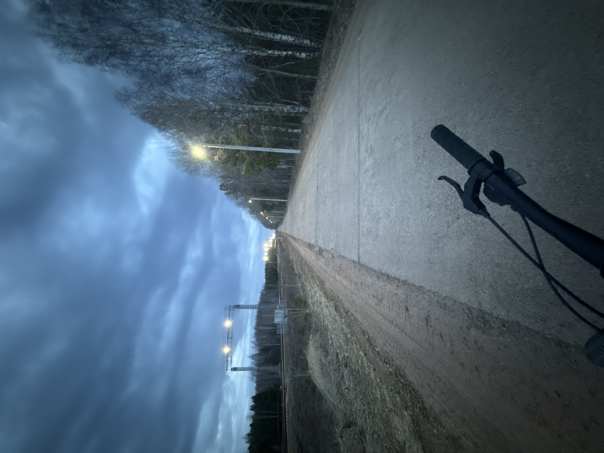 The image depicts a low-angle, first-person perspective of a bicycle ride along a path. The path runs parallel to a railway track, indicated by overhead gantries with lights. A row of streetlights illuminates the path. On the left, a dense tree line is visible, while the sky is overcast with heavy clouds. The bicycle handlebars and brake levers are visible in the foreground. The location is assessed as Kauchuk Stantsiya, Russia, based on recognizable Russian rail infrastructure and the presence of sparse, hardy vegetation. No people are directly visible. The primary action is the implied bicycle ride.
