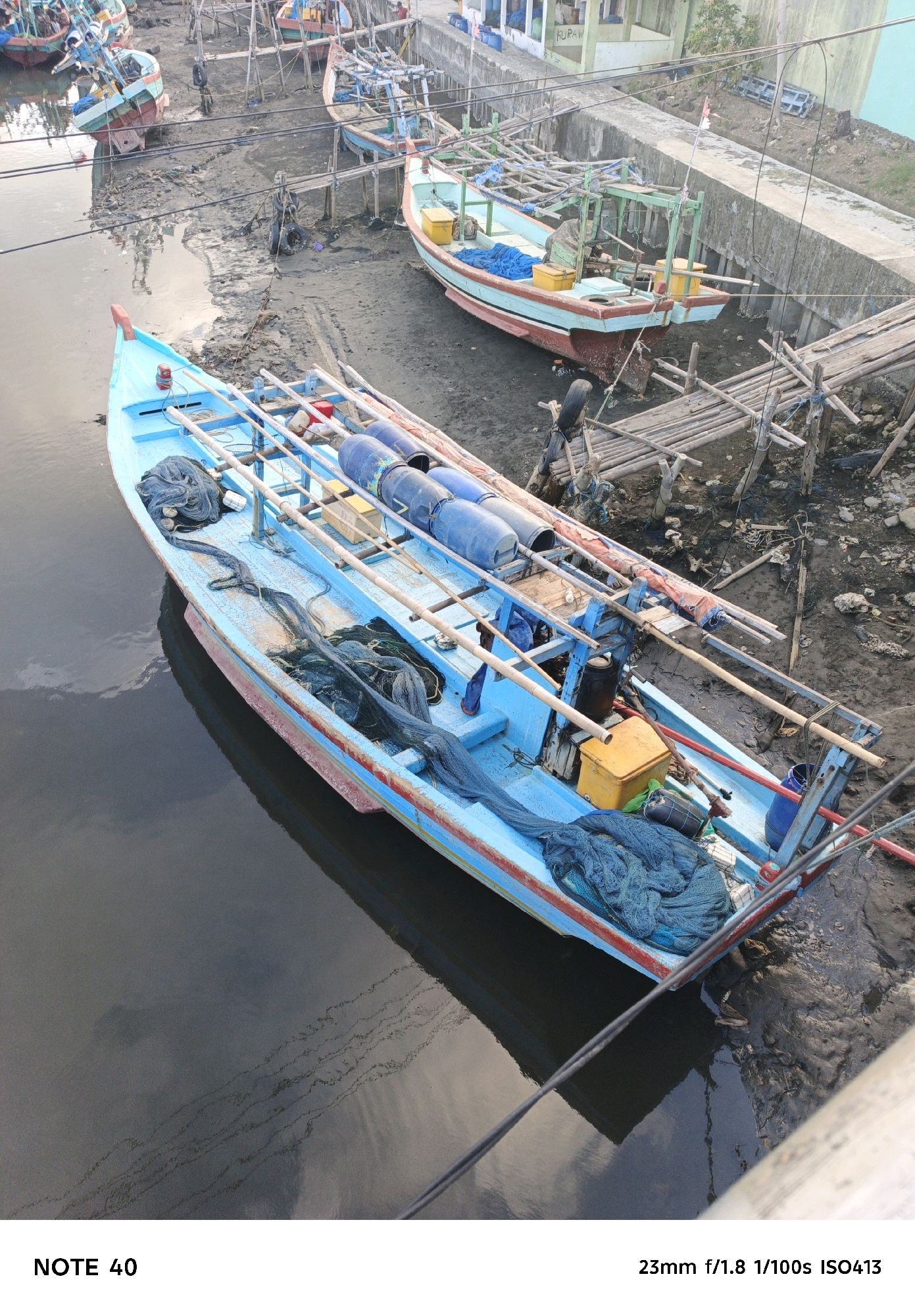 A high-angle shot captures several fishing boats docked along a muddy shoreline in Kelurahan Cilacap, Kecamatan Cilacap, Indonesia. The most prominent vessel is a blue and red fishing boat in the foreground, filled with blue netting and several large blue barrels. To its right, another fishing boat is partially visible, also laden with netting. In the background, more boats are docked, some with colorful hulls. The environment appears to be a canal or a shallow waterway with visible mud and debris along the banks, suggesting a working port or fishing village. The overall scene conveys a sense of everyday activity in a coastal Indonesian community.
