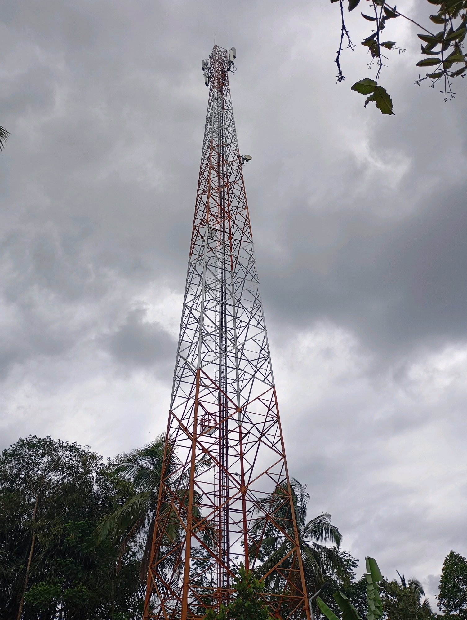 The image shows a tall telecommunications tower against a cloudy sky. The tower is made of a metallic lattice structure, painted with sections of orange and white. Several antennas are mounted at the top of the tower. In the foreground and surrounding the base of the tower, there are trees and lush green foliage. The environment appears to be natural or semi-rural. There are no people or animals visible in the image. The location is identified as Muncang, Indonesia, suggesting an outdoor scene in a tropical or subtropical region given the vegetation.