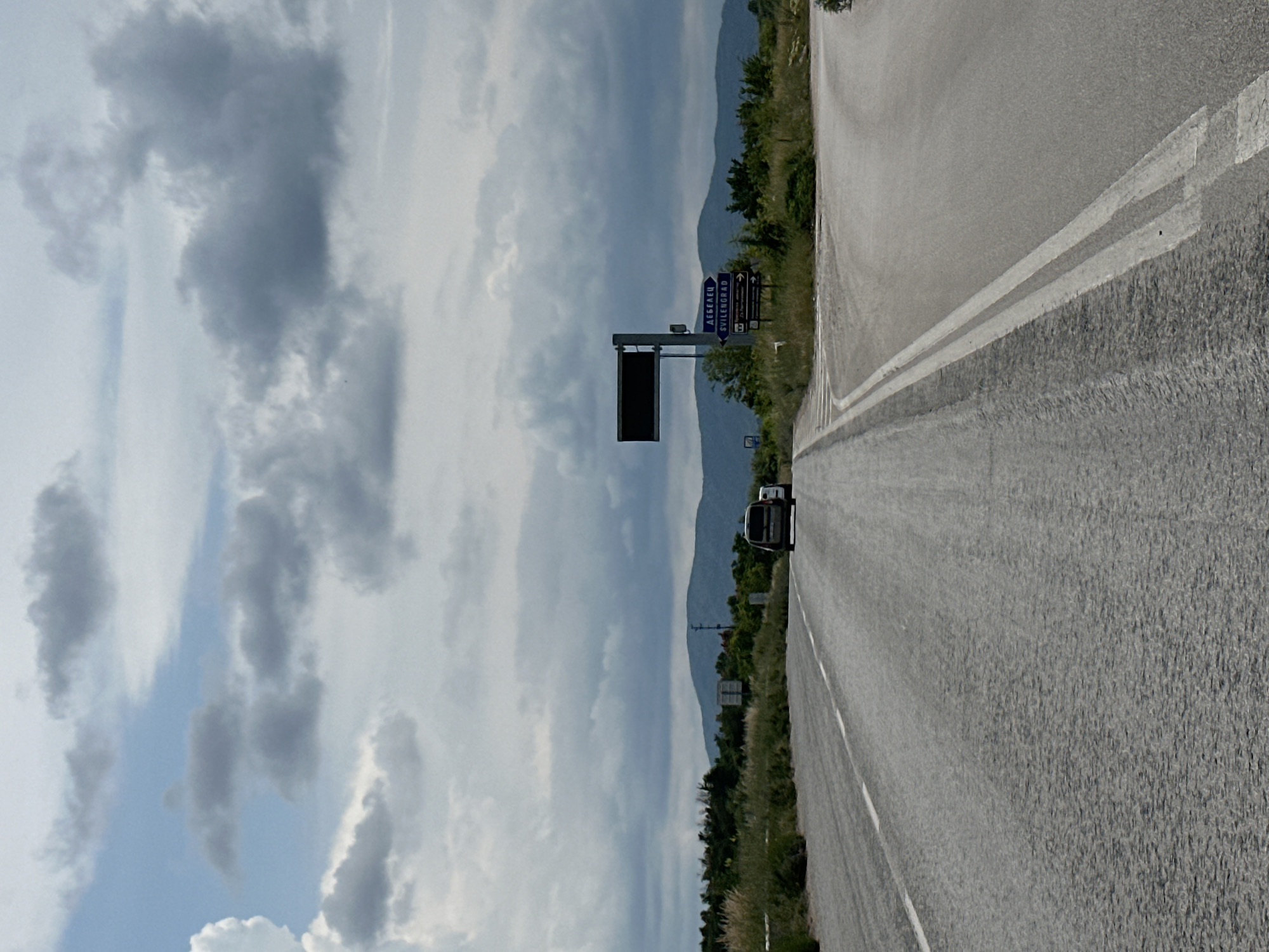 A daytime view captures a stretch of paved road in Prisovo, Bulgaria, under an overcast sky. The road, featuring white solid and dashed lane markings, extends into the distance, gently curving towards the right. A metal guardrail runs along the left edge of the road, which is flanked by green vegetation including bushes and grasses.
In the mid-ground on the left, a large gantry structure holds a dark, blank electronic sign. Below and to its right, a blue directional road sign is mounted on a pole. This sign displays place names in both Cyrillic and Latin script, along with directional arrows. The visible text on the blue sign includes: "АВЕЕЛЕЦ", "СВИЛЕНГРАД" (with "SVILENGRAD" below it), and "ЛЮБИМЕЦ". Further down the road, a white vehicle, possibly a van or SUV, is visible. Rolling mountains or hills form the backdrop, silhouetted against the sky which is filled with prominent grey and white clouds, with patches of lighter blue showing through. The overall scene suggests a journey through a rural or semi-rural landscape.