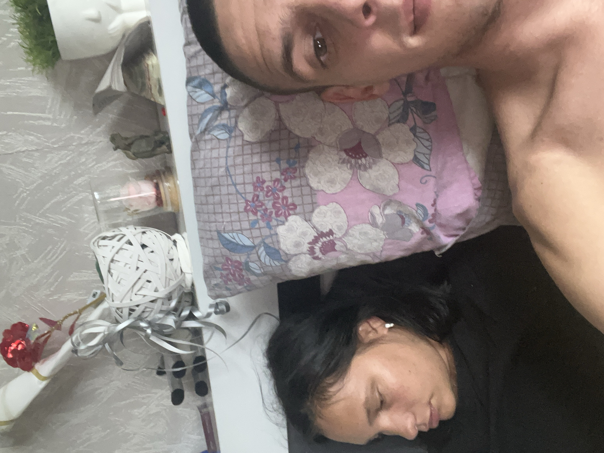 The image captures a close-up, high-angle view of two individuals relaxing, likely in a bedroom. In the foreground, a man with short dark hair and a bare chest lies on a pillow, looking directly at the camera with a neutral expression. The pillow has a floral pattern in shades of pink, purple, white, and blue.
Next to him, partially obscured by the pillow and the man's arm, a woman with dark hair is lying down with her eyes closed, appearing to be asleep or resting deeply. She is wearing a dark top.
To the left of the frame, a side table or shelf is visible against a light grey patterned wall. On this surface, several objects are placed: a small green plant, some papers or books, a clear glass container holding a pink item, a large decorative sphere made of white elastic bands, and a white ornament with a red flower-like top, adorned with silver ribbons.
The scene suggests a moment of intimacy and relaxation, possibly a selfie taken while one person is resting. The lighting appears to be interior, not strongly indicating a specific time of day. There is no visible text in the image. The location is stated to be Sofia, Bulgaria, though no visual cues confirm this.