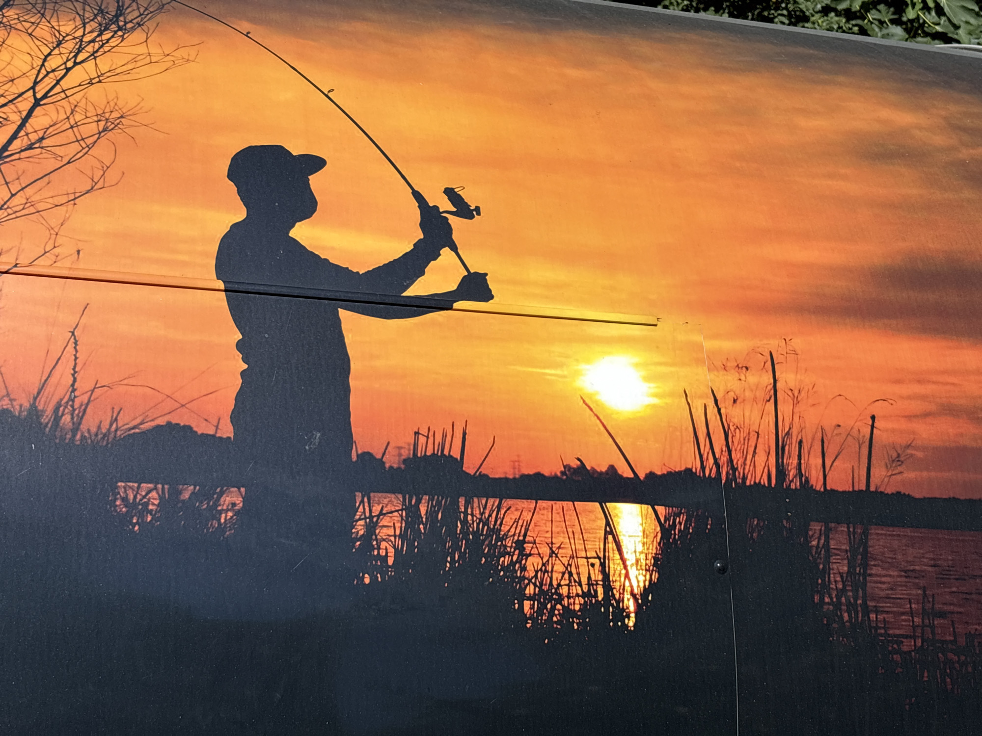 The image is a photograph of a mural or decal depicting a silhouetted fisherman casting a line into a body of water at sunset. The scene is set against a vibrant orange and yellow sky, with the sun low on the horizon casting a strong reflection on the water. Reeds and grasses are silhouetted along the shoreline in the foreground. The mural is applied to a dark, possibly metallic surface, as evidenced by reflections and visible fasteners. There is no indication of the specific location in Plovdiv, Bulgaria, within the image itself.