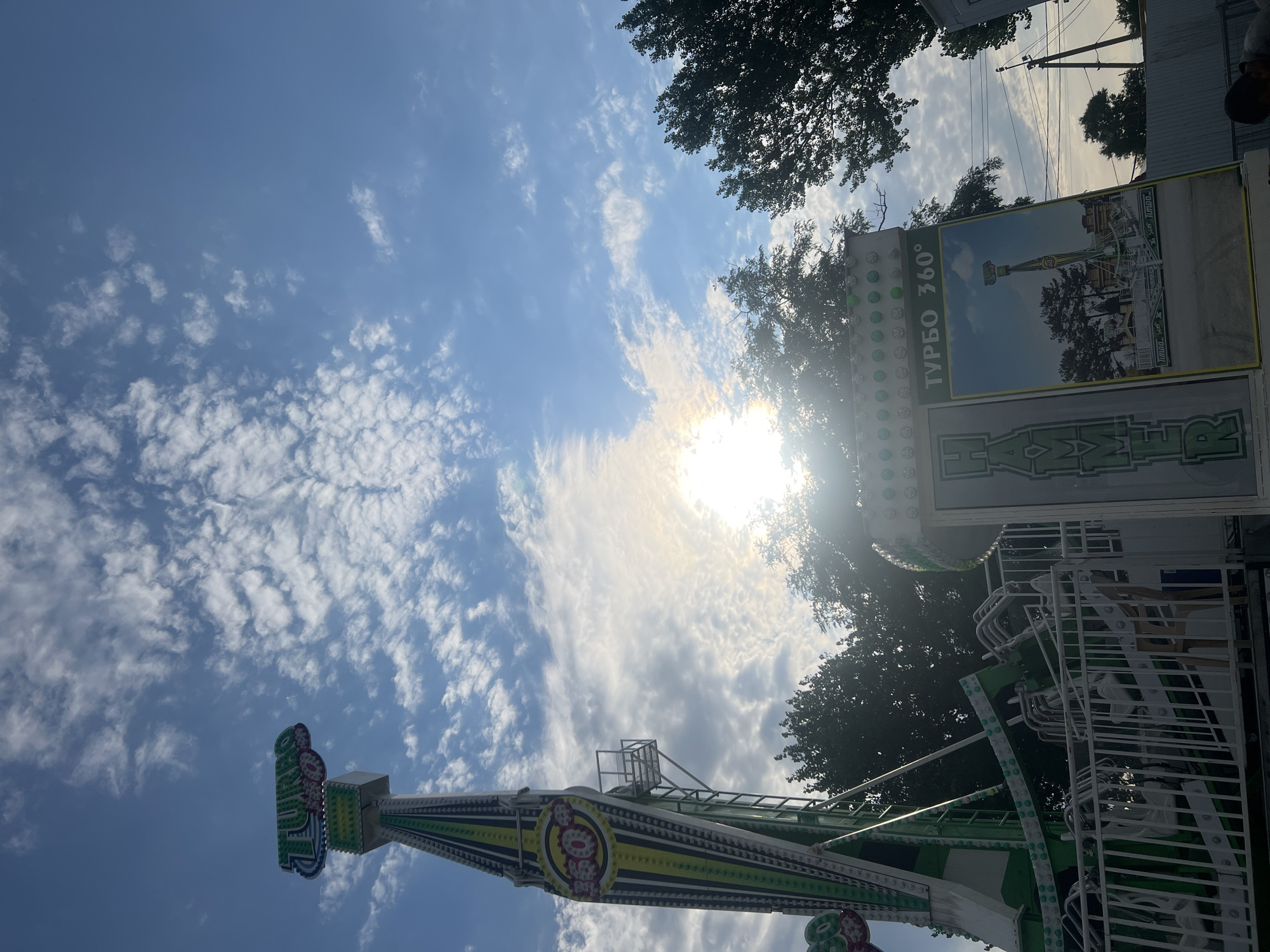 The image captures a portion of an amusement park ride, identified as "Turbo 360," in Golubitskaya, Russia. The ride features a tall, green and white structure with lights and a large "Turbo" sign at the top. In the foreground, a smaller part of the ride is visible, including seats and a metal railing. A large advertisement board for the ride, showing an image of the full ride, is also present. The sky is blue with scattered white clouds, and the sun is shining brightly, creating a lens flare effect. Trees are visible in the background, along with utility poles and a fence. The overall scene depicts a sunny day at an outdoor amusement park.