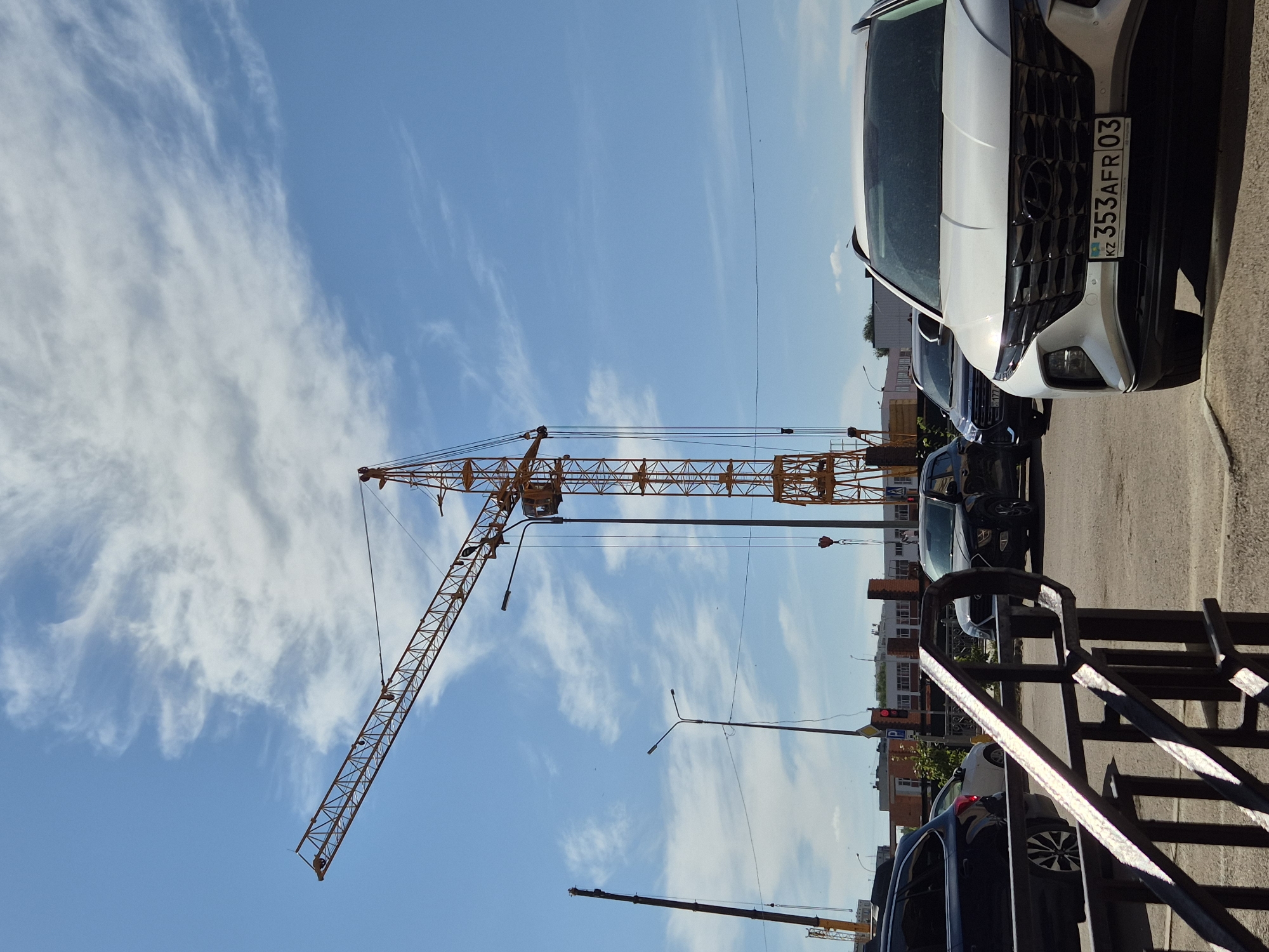This image depicts an outdoor scene in Kokshetau, Kazakhstan, on a bright, sunny day with scattered clouds. The foreground is dominated by a black metal railing. Behind the railing, a white SUV is parked, facing the camera. Its license plate is visible, reading "KZ 353AFR 03". To the left of the white SUV, a black SUV and a white car are parked in a line. Further in the background, a large yellow construction crane with a red hook is prominently featured, along with another, smaller crane to its left. Streetlights are visible, and several multi-story buildings form the backdrop. The scene appears to be a street or parking area adjacent to a construction site.