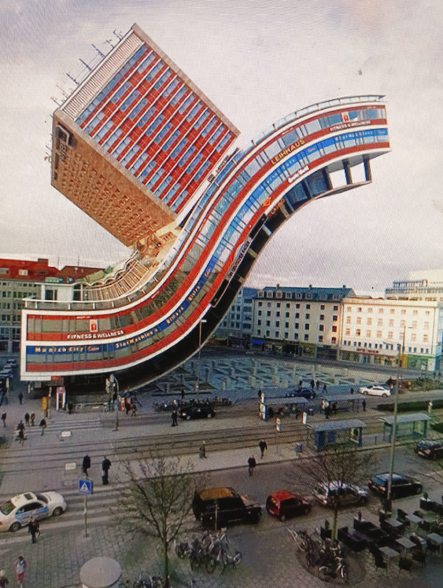 The image depicts the unusual architecture of the "Lehmus House" building in Slutsk, Belarus. The building's design is characterized by its cantilevered upper sections, creating a dynamic, wave-like appearance. The facade features a prominent red, white, and blue color scheme. The ground level of the building is integrated into a street scene, with a pedestrian plaza in front of it. Several people are visible walking in the plaza and on the surrounding sidewalks. Various vehicles, including cars and what appear to be bicycles, are parked or moving in the area. The background shows other urban buildings. The overall environment suggests an active, daytime scene in a city center.