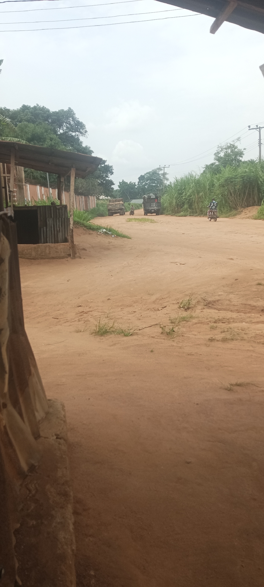 The image depicts a dirt road in Amauzu, Nigeria. Two trucks and a motorcycle are visible on the road. Vegetation lines the sides of the road. A building with a wooden structure is on the left. The sky is overcast.