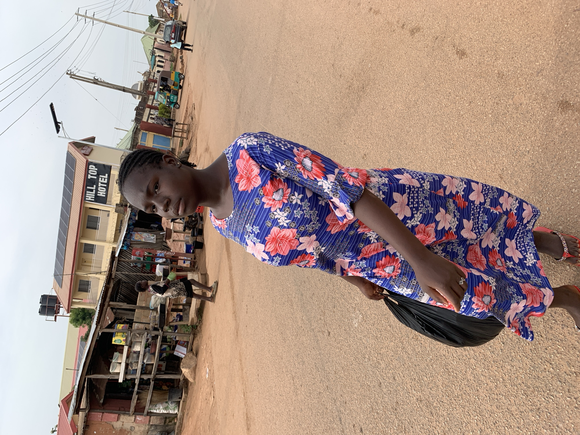 A young woman with braided hair and dark skin walks down a dusty, unpaved street in Jos, Nigeria. She wears a blue dress with a floral pattern of red and pink flowers. Her left hand holds a large black plastic bag, and her right hand rests on her hip. She is wearing red flip-flops with jeweled straps. In the background, several buildings line the street, including a two-story building with "HILL TOP HOTEL" written on its facade and solar panels on the roof. A few shops with open stalls are visible to the left, displaying various goods. Several people and vehicles, including a small auto-rickshaw and a car, are present in the background. The sky is overcast.