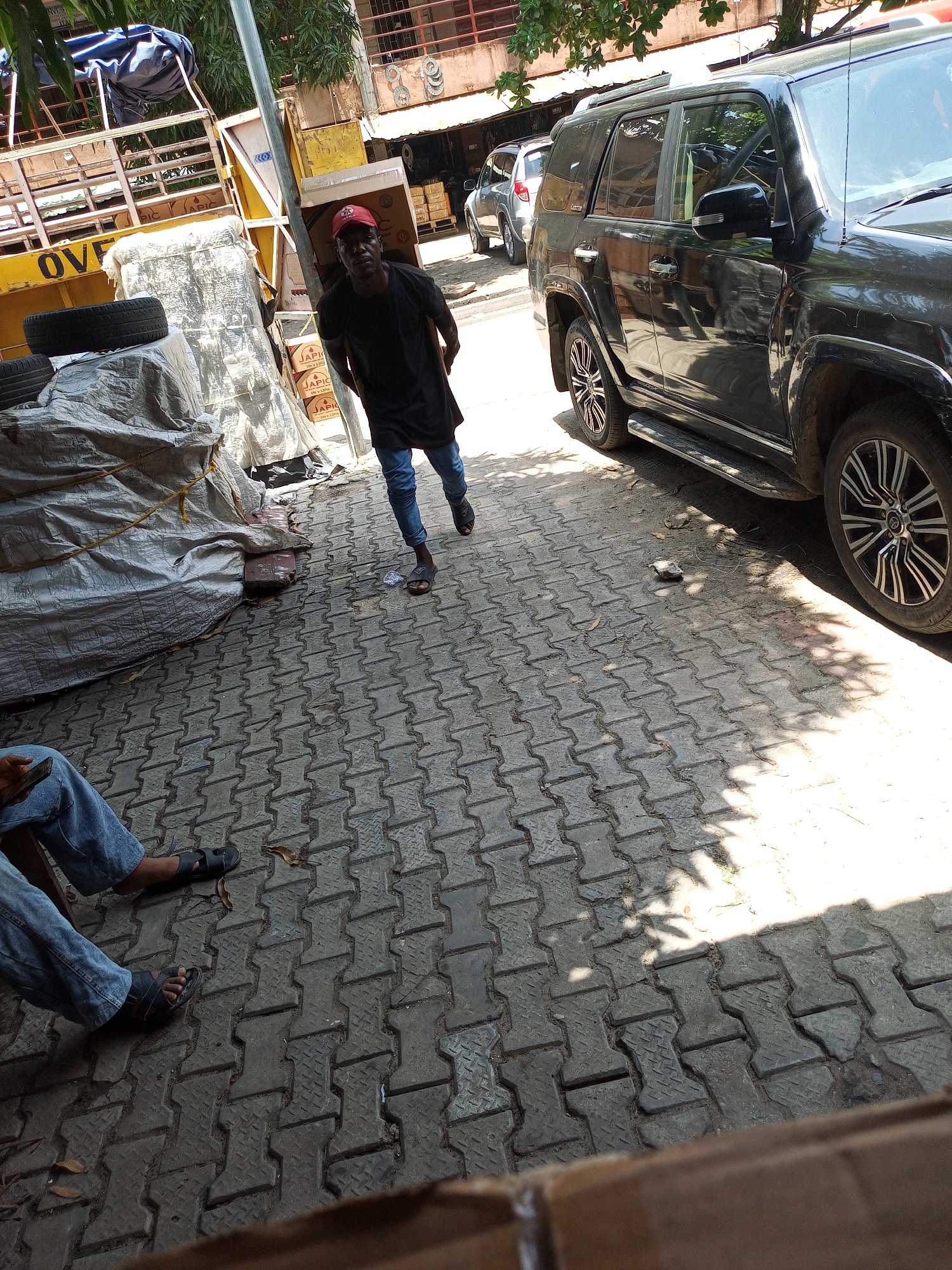 The image captures an outdoor urban scene, likely in Satellite Town, Nigeria, under bright midday sun. The ground is paved with distinctive gray interlocking concrete blocks, and dappled shadows from trees indicate clear weather.
In the mid-ground, a dark-skinned man walks towards the camera. He wears a black t-shirt, blue jeans, and a red baseball cap, with a neutral expression. To his right, a black SUV, resembling a Toyota 4Runner, is parked on the paved area, featuring roof rails and stylized black and silver wheel rims.
The left side of the frame is dominated by a large, yellow truck or trailer structure, with the word "OVER" visible on its side. Adjacent to it, various items are covered with gray tarpaulins, and two car tires are stacked on one. Cardboard boxes labeled "JAPIC" and "10's x 10PCS" are visible near the covered goods, suggesting commercial activity.
In the bottom-left corner, the legs and feet of another person are visible, clad in blue jeans and dark sandals. This individual appears to be seated or resting, holding a smartphone. Buildings with visible upper levels and green foliage from trees are present in the background, completing the bustling street setting.
