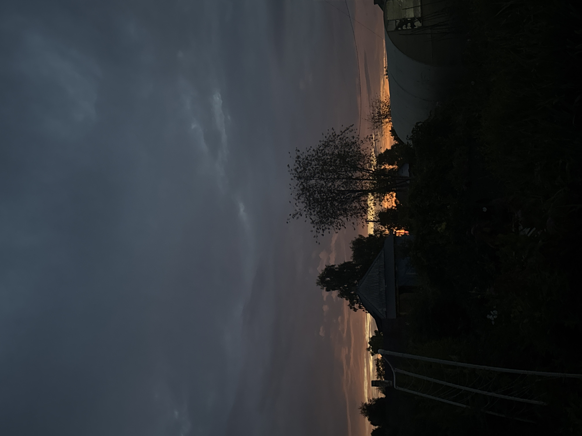 The image depicts a rural scene at dusk in Ryabinino, Russia. The sky is overcast with dark gray clouds, but a band of orange and yellow light from the setting sun is visible near the horizon, peeking through gaps in the clouds. In the foreground, silhouettes of dense vegetation and trees are prominent, obscuring much of the ground.
To the left, a white, angled structure, possibly a sports goal or frame, is partially visible. In the mid-ground, a small, weathered building with a dark, corrugated roof and visible dark window openings is situated among the trees. To the right of the building, a large, arched greenhouse structure made of translucent material is present. The overall impression is one of twilight in a semi-natural, semi-cultivated environment. There are no people or animals visible in the image.