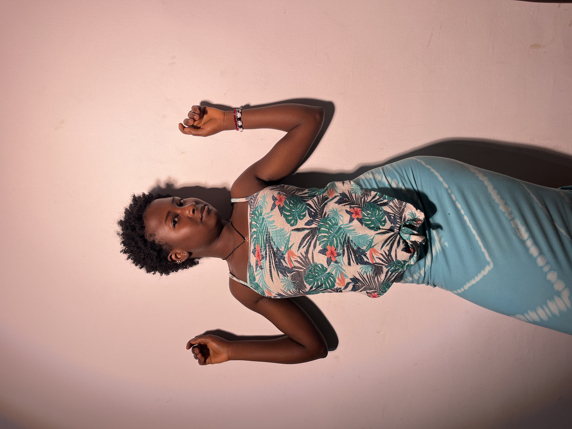 In an indoor setting with a light pink, textured wall, a young woman with dark, curly hair stands with her arms bent at the elbows, hands raised and fists clenched. She is wearing a light-colored tank top with a green, white, and pink floral pattern, and a light blue skirt with a white tie-dye pattern. A dark necklace with a pendant is visible around her neck, and a bracelet with red and white beads is on her right wrist. The lighting creates a shadow of her on the wall to her left. The background is plain and uncluttered, suggesting an indoor space.