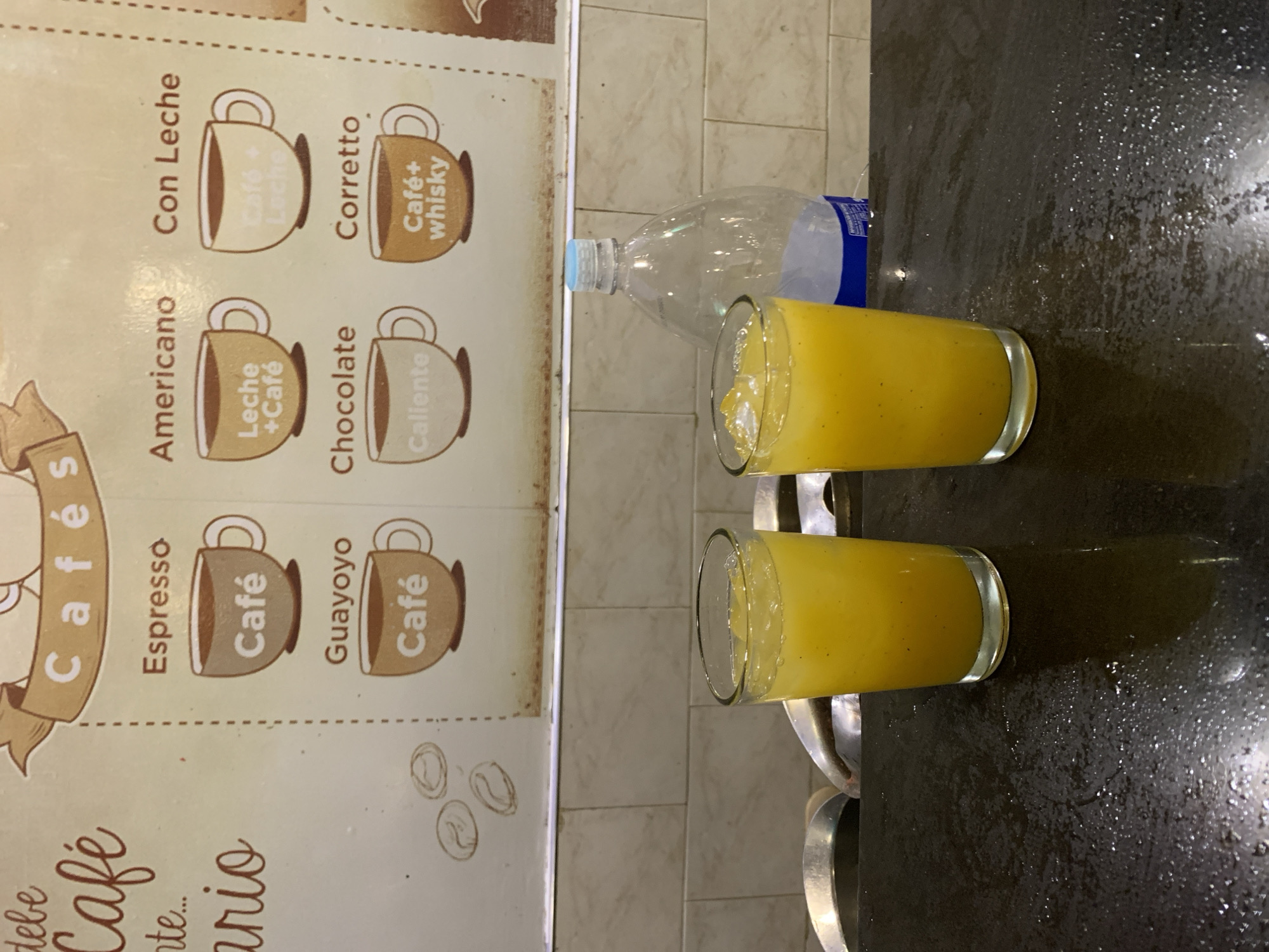 This image depicts an indoor scene in Playa El Agua, Venezuela, featuring a coffee menu sign above a counter with two beverages.
The upper portion of the image displays a light-colored sign, likely a menu board, with "Cafés" written prominently at the top center. Below this, a grid of six coffee options is illustrated with cartoon coffee cups and text. These options include: "Espresso" (labeled "Café"), "Americano" (labeled "Leche + Café"), "Con Leche" (labeled with faded "Café + Leche"), "Guayoyo" (labeled "Café"), "Chocolate" (labeled "Caliente"), and "Corretto" (labeled "Café + whisky"). To the left of the coffee options, partial text reads "debe Café nte..." and "ario," accompanied by illustrations of coffee beans. The sign shows some discoloration or wear along its lower edge.
Below the sign, a light-beige tiled wall is visible, with rectangular tiles arranged in a horizontal grid pattern. A thin metallic strip runs horizontally between the sign and the tiles.
The lower portion of the image features a dark, reflective counter surface, exhibiting numerous water droplets or condensation marks. On this counter, two clear glasses are placed, each containing a vibrant yellow-orange, pulpy liquid with ice cubes. Small, dark specks are visible within the liquid. Behind the rightmost glass, a tall, clear plastic bottle with a light blue cap and a blue label at its base stands upright; it appears mostly empty. In the background, behind the glasses and bottle, sections of curved, metallic objects are partially visible, suggesting kitchen or bar equipment.
No individuals are present in the image.