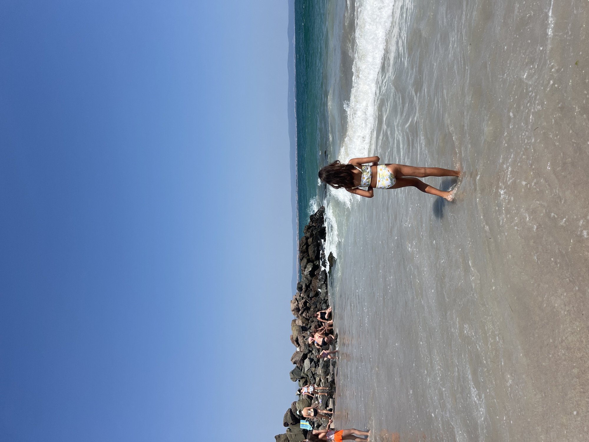 The media file depicts a bright, sunny day at a beach in Pomorie, Bulgaria. The scene is dominated by a clear blue sky on the left and the ocean and shoreline on the right. A young girl with long, dark hair, wearing a yellow and white patterned bikini, is standing in shallow water. Her back is to the camera as she looks out towards the waves.
In the background, a jetty made of dark, rough rocks stretches into the sea, with several people sitting and lounging on it. These individuals are also dressed in swimwear, suggesting they are enjoying a day at the beach. The waves are gentle, lapping at the shore and creating a tranquil atmosphere. The sand is wet and reflective from the water. The sun appears to be high in the sky, casting shadows and indicating it is likely midday. The overall impression is one of leisure and a typical summer day at the coast. There is no visible text in the image.