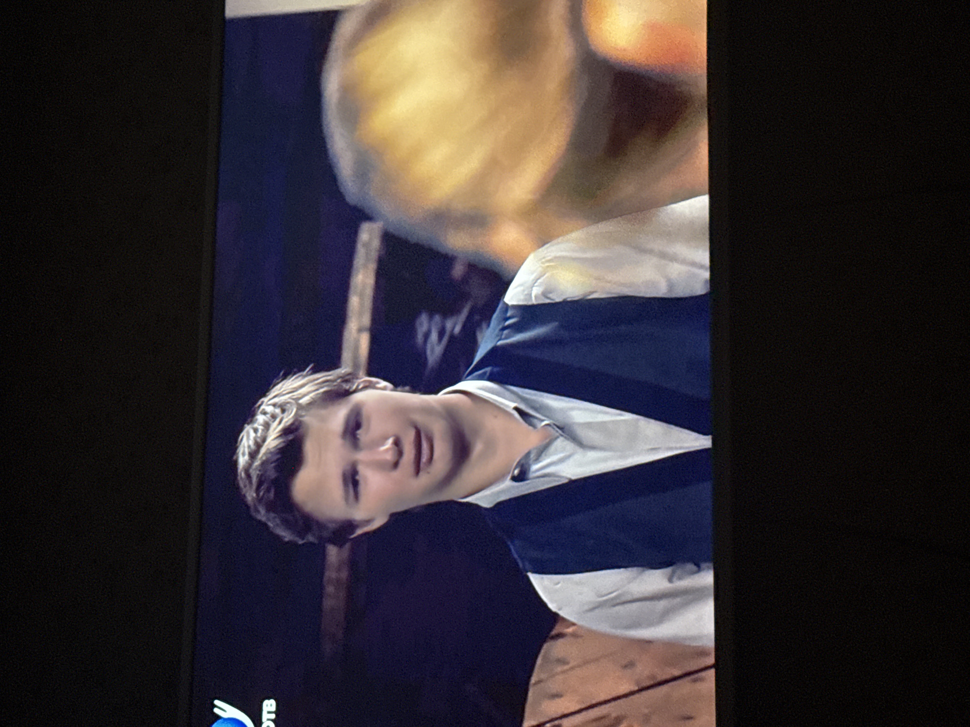 A medium shot captures a man wearing a white collared shirt with a navy blue vest. The man appears to be the focus of a television screen. Part of another person’s face is visible in the upper right corner of the image. The bottom left corner displays the logo for "DTB," a Russian television channel. The background is dark and blurred, possibly indicating an indoor setting. The image does not contain sufficient information to discern the city of Mytishchi, Russia.