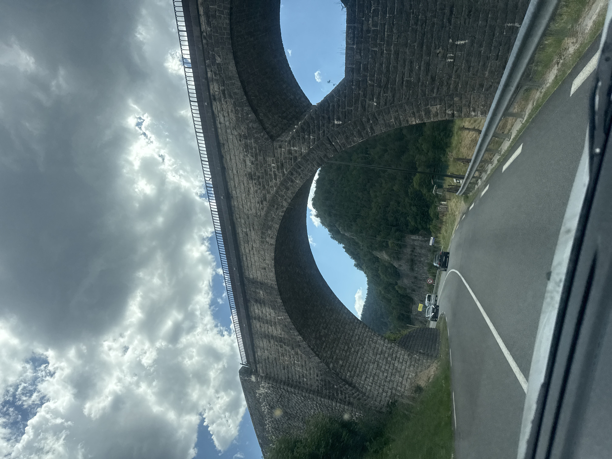 The image captures a low-angle view of a large stone viaduct, likely a railway bridge, with its arches framing a lush, green mountainside and a bright blue sky interspersed with white, fluffy clouds. The stone construction of the viaduct is detailed, showing individual blocks and the curved architecture. A modern metal guardrail runs alongside a paved road visible to the right of the frame, indicating a route passing beneath or near the viaduct. Several cars are seen on the road, suggesting ongoing traffic. The overall scene is bathed in daylight, with the sun creating bright patches on the clouds and illuminating the landscape. The orientation of the image is rotated, making it appear as if the viewer is looking upwards from ground level or from a vehicle driving along the road. The setting appears to be a natural, possibly mountainous area, given the dense vegetation on the slopes. No people are directly visible, but their presence is implied by the cars on the road. The time of day appears to be daytime, likely late morning or afternoon due to the bright sunlight and cloud cover. There is a speed limit sign visible on the side of the road with "60" indicating kilometers per hour. The location is identified as Hauts de Bienne, France.