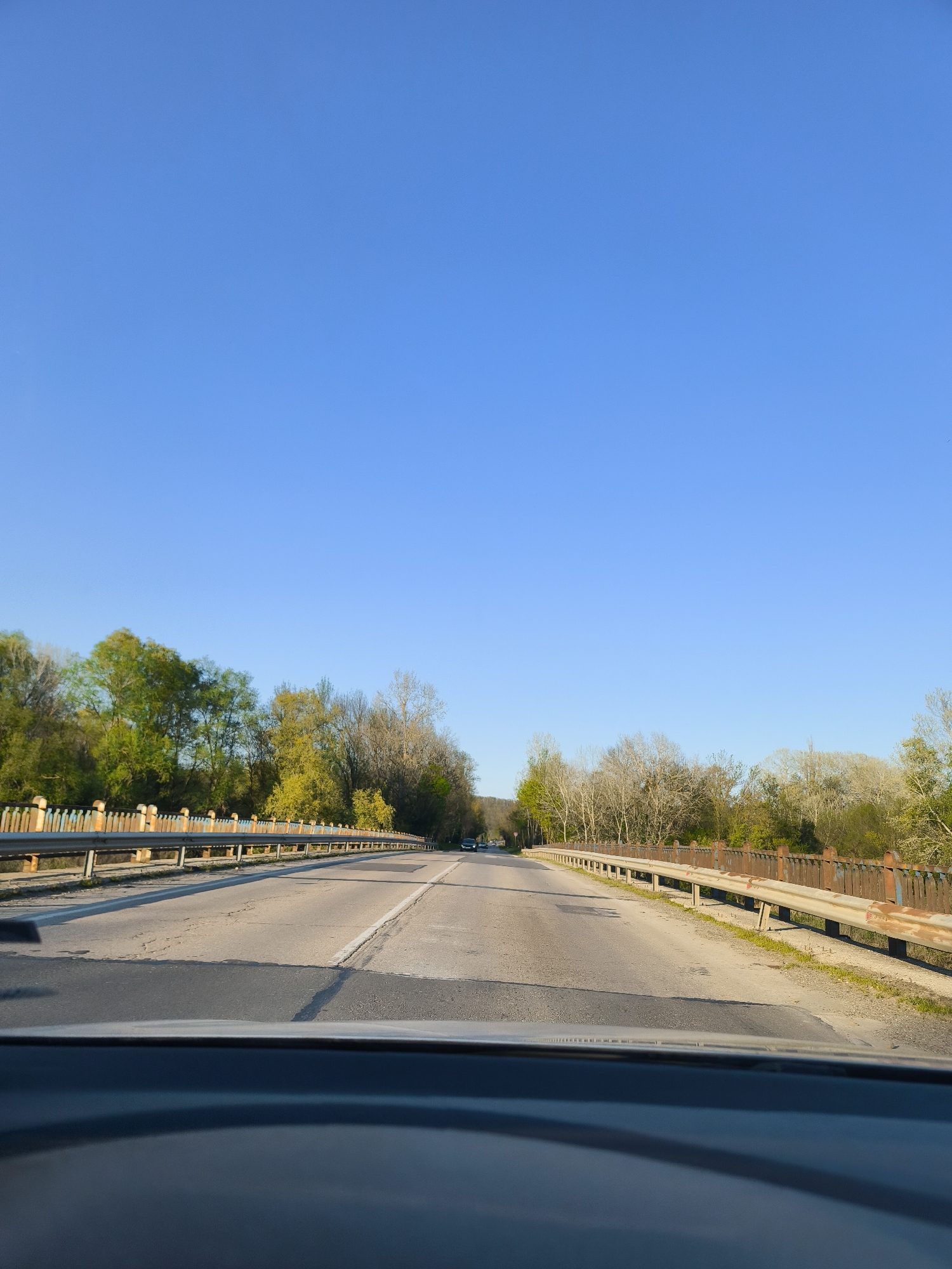 A forward-facing view from inside a car traveling on a bridge near Staro Oriahovo, Bulgaria. The road is asphalt and has a white line dividing the lanes. The bridge has old, weathered metal railings on both sides. Trees line both sides of the road, showing early spring foliage. The sky is clear blue. A car is visible in the distance ahead. The dashboard of the car taking the photo is visible in the foreground.