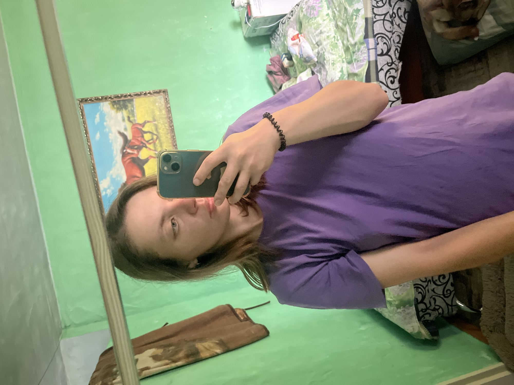 A young woman with brown hair is taking a mirror selfie in a room with bright green walls. She is wearing a purple t-shirt and a black spiral bracelet on her left wrist. She is holding a dark-colored smartphone in her right hand, partially obscuring her face. In the reflection, a framed picture of two horses is visible on the wall behind her. To the right of the picture, part of a bed with a patterned duvet is visible. A brown towel or blanket hangs on the wall to the left of the frame. The overall impression is an informal, personal snapshot taken in a residential setting. The image was taken in Nizhny Tagil, Russia.