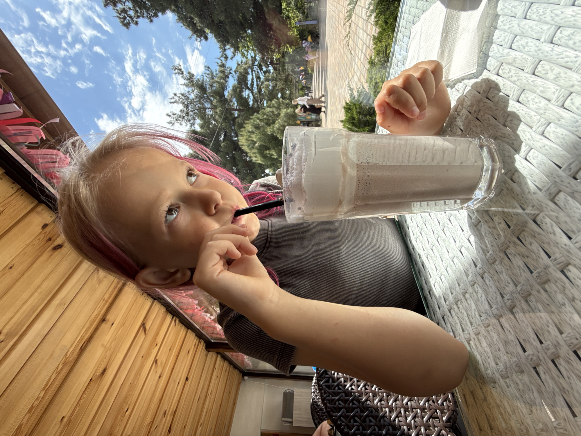 A low-angle, portrait-oriented shot captures a young child enjoying a milkshake at an outdoor cafe. The child, with fair skin, light-colored hair featuring vibrant pink streaks, and blue eyes, is positioned in the lower-left of the frame, looking upwards and slightly to the right. They wear a dark grey, ribbed long-sleeved top.
In the center-right, a tall glass of chocolate milkshake with a thick layer of white foam on top is held by the child, who is drinking through a black straw. The child's right hand is visible, resting on a white, woven-textured table with a clear glass top, suggesting an outdoor patio setting.
The background reveals a bright outdoor scene. Lush green trees and foliage are visible under a vivid blue sky adorned with scattered wispy white clouds. A paved pedestrian path stretches into the distance, with a few blurry figures walking, indicating a public park or walkway adjacent to the cafe. To the left, a vertical wooden slatted wall forms part of the cafe's structure, with some colorful, blurred decorative elements hanging above it.
The scene is well-lit by natural sunlight, casting clear shadows on the white woven table, suggesting it is daytime, likely mid-day or afternoon. The child appears calm and focused on their drink. No visible text is present in the image.