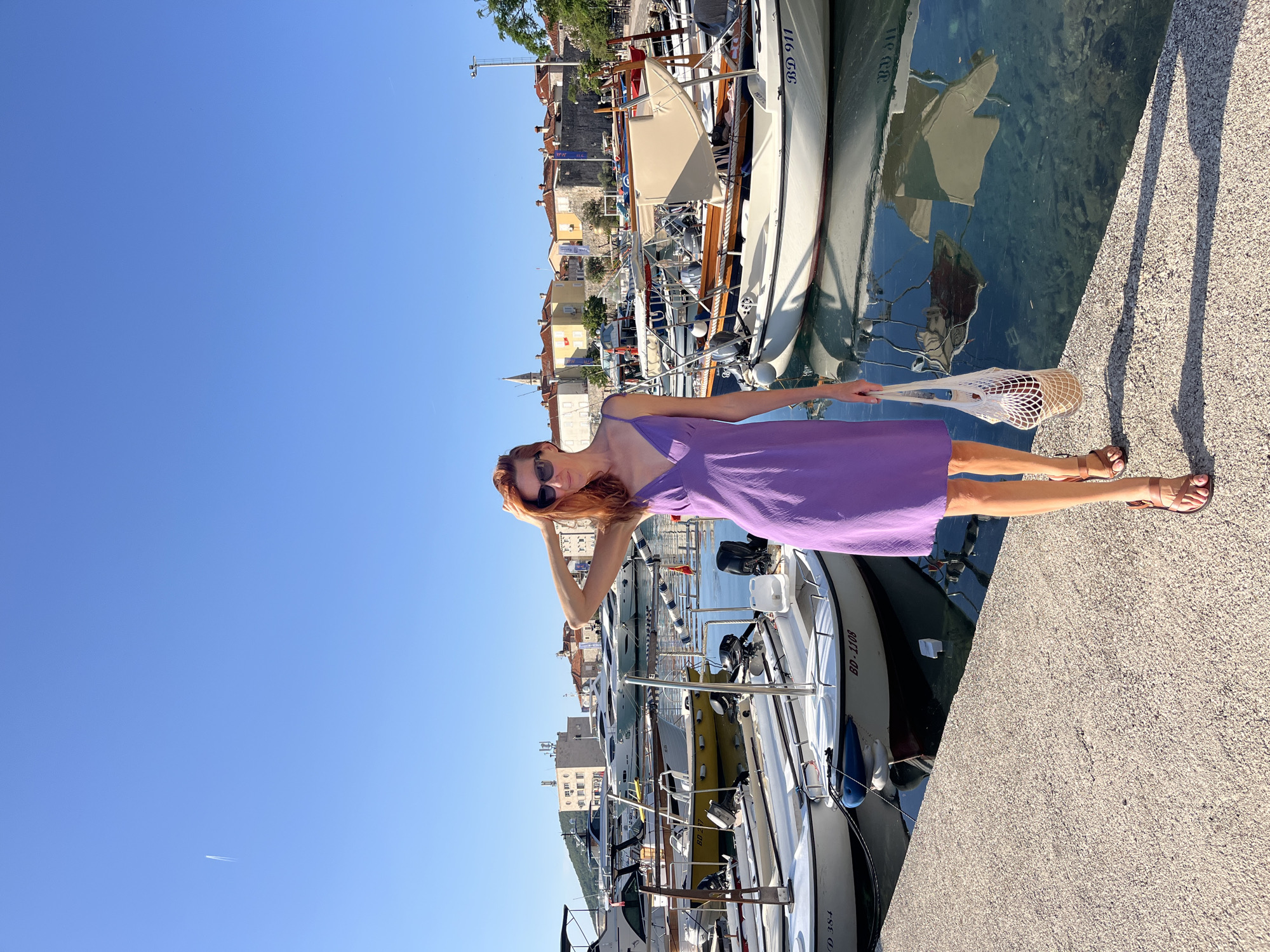 A full-body shot captures a woman standing on a concrete pier at the marina in Budva, Montenegro, under a clear, bright blue sky with a single aircraft contrail in the upper left. The woman, positioned slightly right of center, has reddish-brown hair, wears dark sunglasses, a knee-length purple slip dress, and brown sandals. Her right hand is raised to her head, while her left hand holds a white mesh string bag containing a dark bottle. Her shadow is cast distinctly on the pier to her left.
The pier borders a body of water to her left and behind, which is filled with numerous docked vessels. These include smaller white motorboats with outboards displaying registration numbers like "BD-1108," "BD-384," and "BD 911," as well as larger, luxury yachts further in the background. The water reflects the boats and the sky. The background reveals the cityscape of Budva, characterized by densely packed buildings with red-tiled roofs, yellow and white facades, and a prominent church steeple, all nestled against distant hills. The scene is brightly illuminated by direct sunlight.