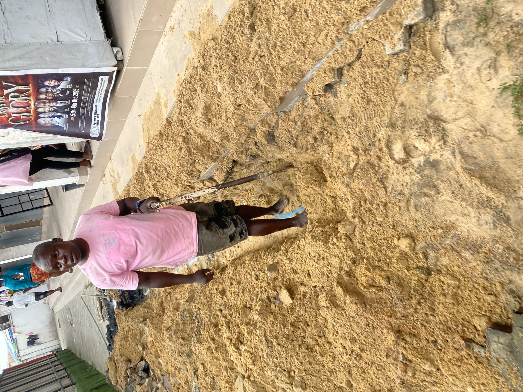 An outdoor scene in Ado, Nigeria, depicts a man engaged in manual labor on a construction or excavation site, dominated by a large mound of sandy earth. The main subject, a man of African descent with a bald head, wears a pink t-shirt with a light grey graphic and dark shorts. He stands on the sandy ground, holding a metal tool resembling a shovel or digging stick, and looks directly at the camera with a neutral expression. His feet are bare.
In the background, several other individuals are partially visible on a paved or tiled area, with one person in a light blue top and another in white. Various debris, small rocks, and metal bars are scattered within the sand mound, suggesting ongoing work. To the far left, a vertical banner is affixed to a structure, advertising an event.
The banner prominently displays the text "DAYS OF GLORY" and the dates "5th - 7th Oct. 2025." It also features portraits of several individuals and mentions "POTTER'S TABERNACLE MINISTRIES." Smaller legible text includes names like "BISHOP ISAAC," "PASTOR CHRIS," "PST MRS KUNLE," "PASTOR YINKA," and "EVANG. OLORI," along with "HOST PASTOR."
The lighting suggests a clear daytime, likely morning or afternoon. The overall setting is consistent with an urban or semi-urban environment undergoing development or renovation.