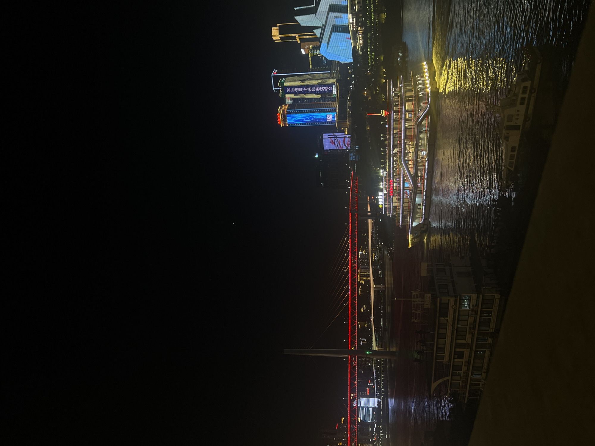 This image captures a nighttime view of the Yuzhong District in Chongqing, China. The scene is dominated by the brightly lit skyline featuring modern skyscrapers with illuminated facades and signage. A prominent red-lit bridge spans across a body of water, likely the Yangtze River, with traffic visible on its span. In the foreground, a large, multi-decked tourist boat with numerous lit windows and balconies is navigating the water, its lights reflecting on the dark surface. Another smaller vessel is moored or moving slowly nearby. The overall atmosphere is that of a vibrant, urban waterfront at night.