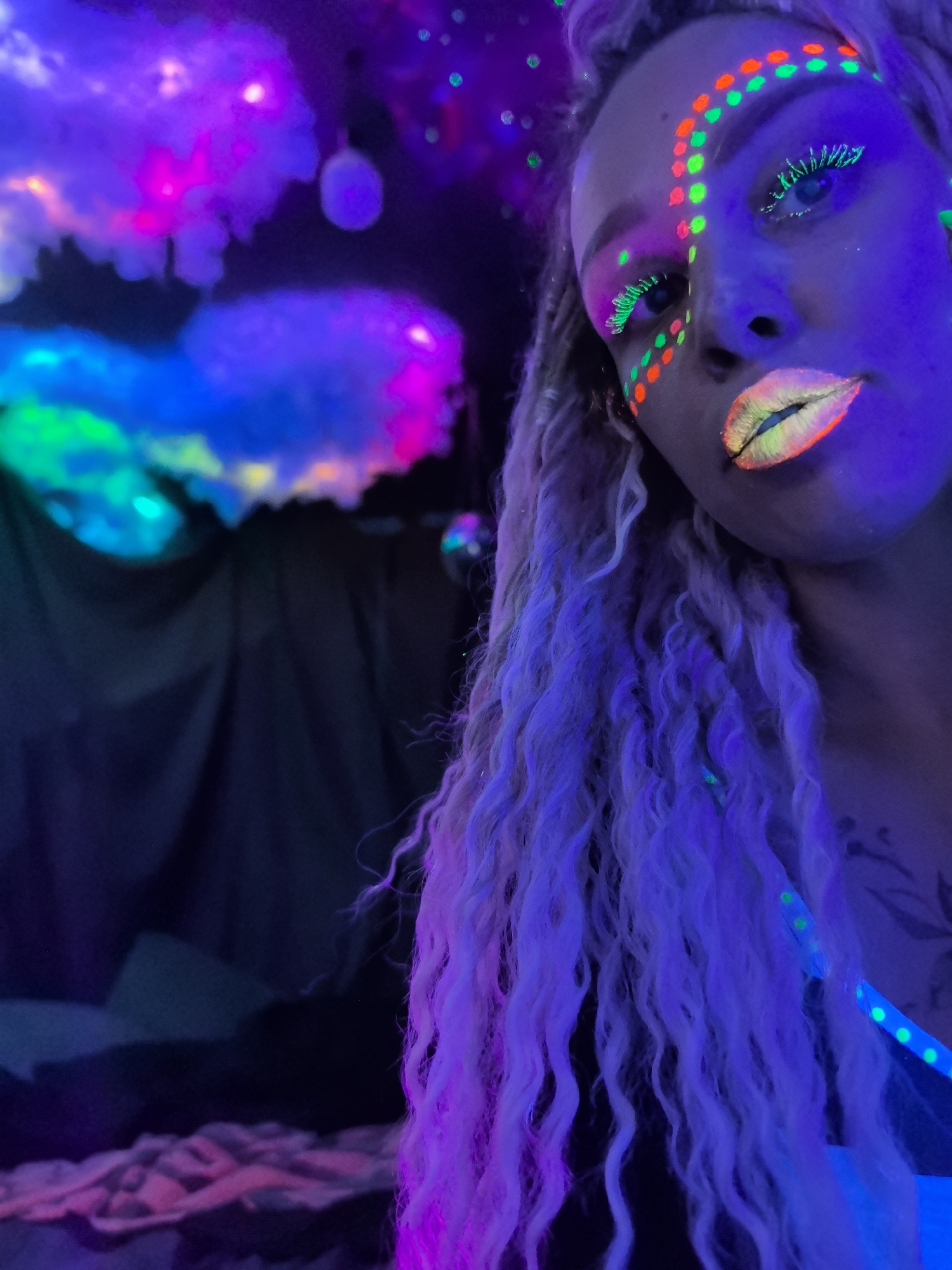 A close-up, angled portrait of a fair-skinned person with blonde, curly hair illuminated under blacklight. The person has bright makeup including green-and-orange dots leading from the hairline down the cheekbones, bright green eyelashes, and gradient lips (yellow-green in the middle, transitioning to orange at the edges). The person is against a backdrop including what appear to be faux clouds illuminated in blue, green, and purple light. The background also includes what appears to be a dark draped fabric and a bed-like surface. The tattoo on the person's upper chest is visible. The scene does not appear to be in Noviy, Russia.