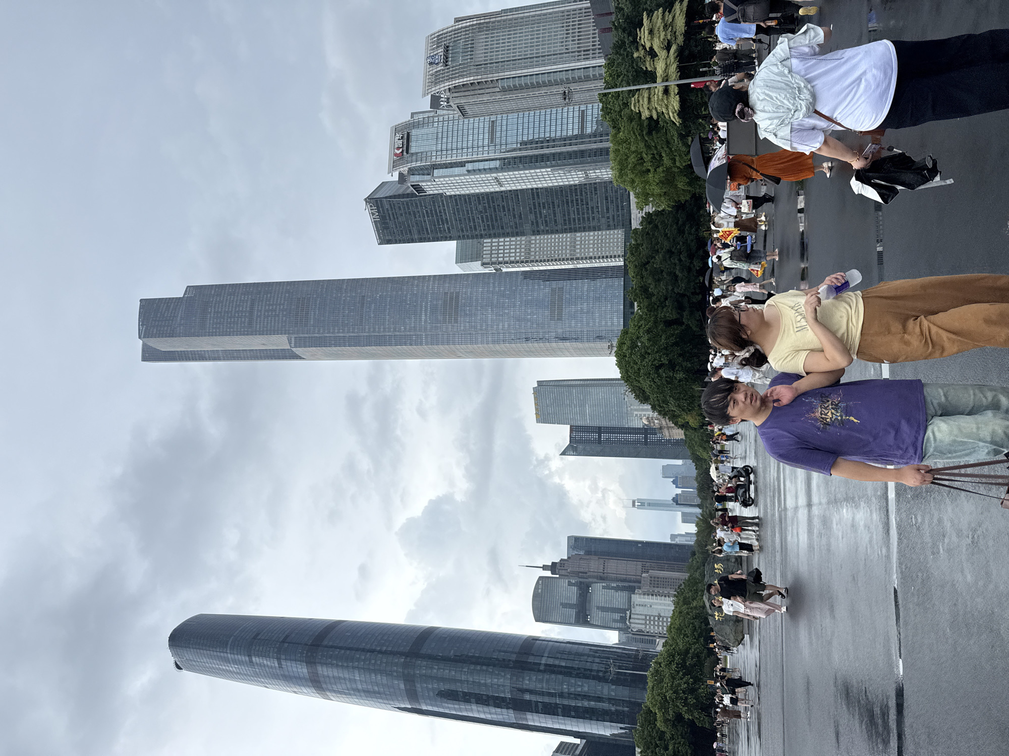 This image depicts a wide, wet, paved plaza in front of a modern cityscape with several tall skyscrapers. The ground is reflective, indicating recent rain. Numerous people are walking across the plaza, some with umbrellas, suggesting it is overcast or still raining lightly. In the foreground, a young man in a purple t-shirt and light-colored pants walks towards the viewer, carrying a tote bag. To his left, a young woman in a yellow t-shirt and brown pants walks away from the viewer, holding a water bottle. Behind them, more pedestrians are visible, including a woman in an orange dress holding an umbrella. The background is dominated by the towering glass and steel structures of a modern business district. One particularly tall, curved skyscraper is prominent on the left side of the image, while two very tall, rectilinear skyscrapers are centrally located. Other modern buildings of varying heights and architectural styles fill the background, interspersed with green trees. The sky is overcast and grey. The scene appears to be a public area within the Tianhe District of China, likely a commercial or financial center.