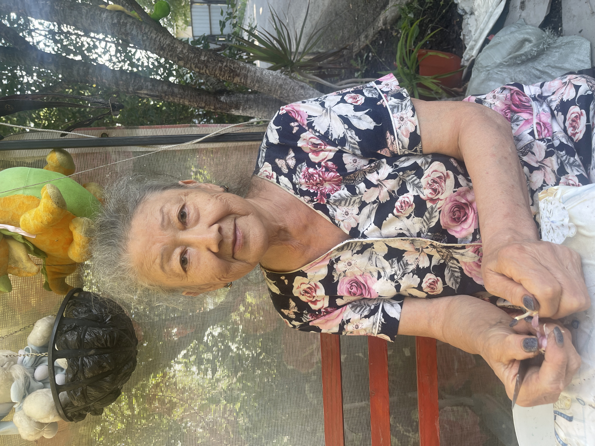 An elderly East Asian woman with graying hair and wrinkles sits on a red wooden bench, facing forward and looking at the camera. She is wearing a dark floral-print pajama top with pink and white roses. Her hands are clasped in her lap, and she is holding a small object and a thin metal tool, appearing to be trimming something. Behind her, a screened porch or greenhouse area is visible, with a hanging black basket filled with black plastic bags. To the right, a tree trunk and branches with some green foliage are seen. The background suggests an outdoor setting with trees and a paved area, possibly a street or driveway. The overall lighting indicates daylight.