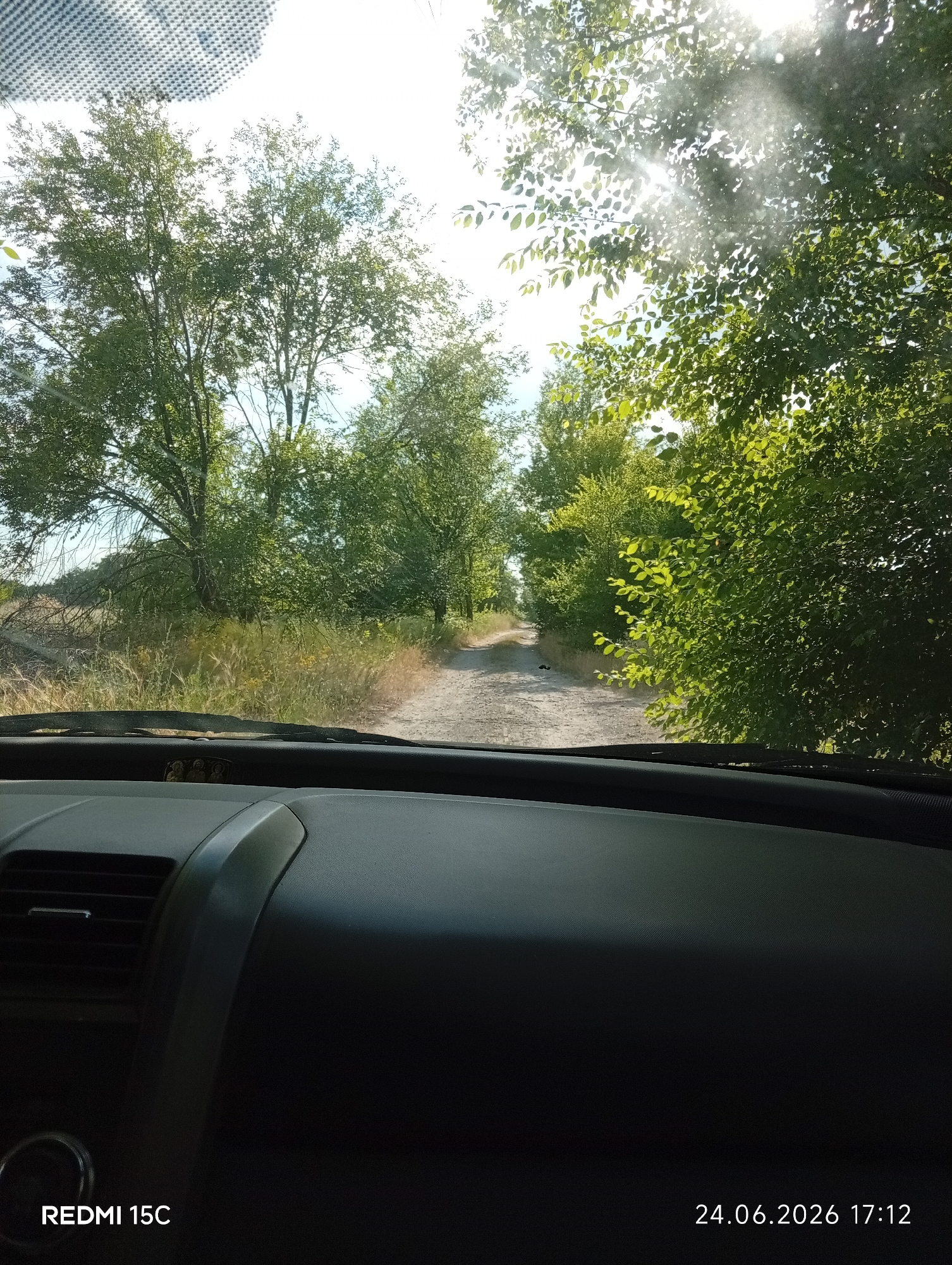 The image captures a view from inside a vehicle, looking out through the windshield. The scene is a dirt road winding through a landscape dominated by lush green trees and foliage. Tall trees line both sides of the road, with some branches extending over the path. Sunlight filters through the leaves, creating a dappled effect on the road and surrounding vegetation. The sky is bright, indicating daytime, and the sun's rays are visible, suggesting clear weather. The road appears to be in a rural or semi-rural area, possibly a natural setting.
The interior of the vehicle is visible in the foreground, with a dark dashboard and air vents. The overall impression is of a scenic drive through a wooded area on a sunny day.
Visible text in the image includes "REDMI 15C" in the bottom left corner, indicating the device used to capture the photo, and a date and time stamp "24.06.2026 17:12" in the bottom right corner. The location context provided is Mykolaivka, Ukraine.
