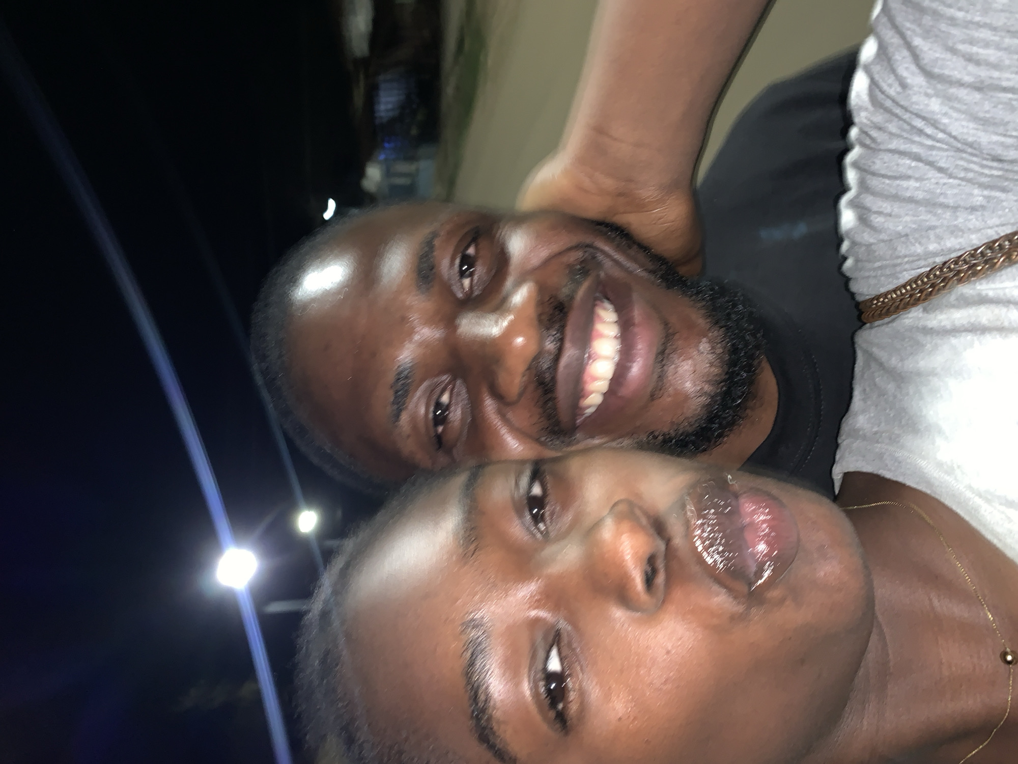 Two people pose closely for a selfie. The man, with dark skin, a shaved head, and a black beard, smiles, showing his teeth. He wears a black shirt. The woman has dark skin and wears a gold necklace. Her lips appear glossy. They are outdoors at night, as evidenced by the dark background and streetlights in the distance. There is no indication that this scene is located in Kabong, Nigeria.