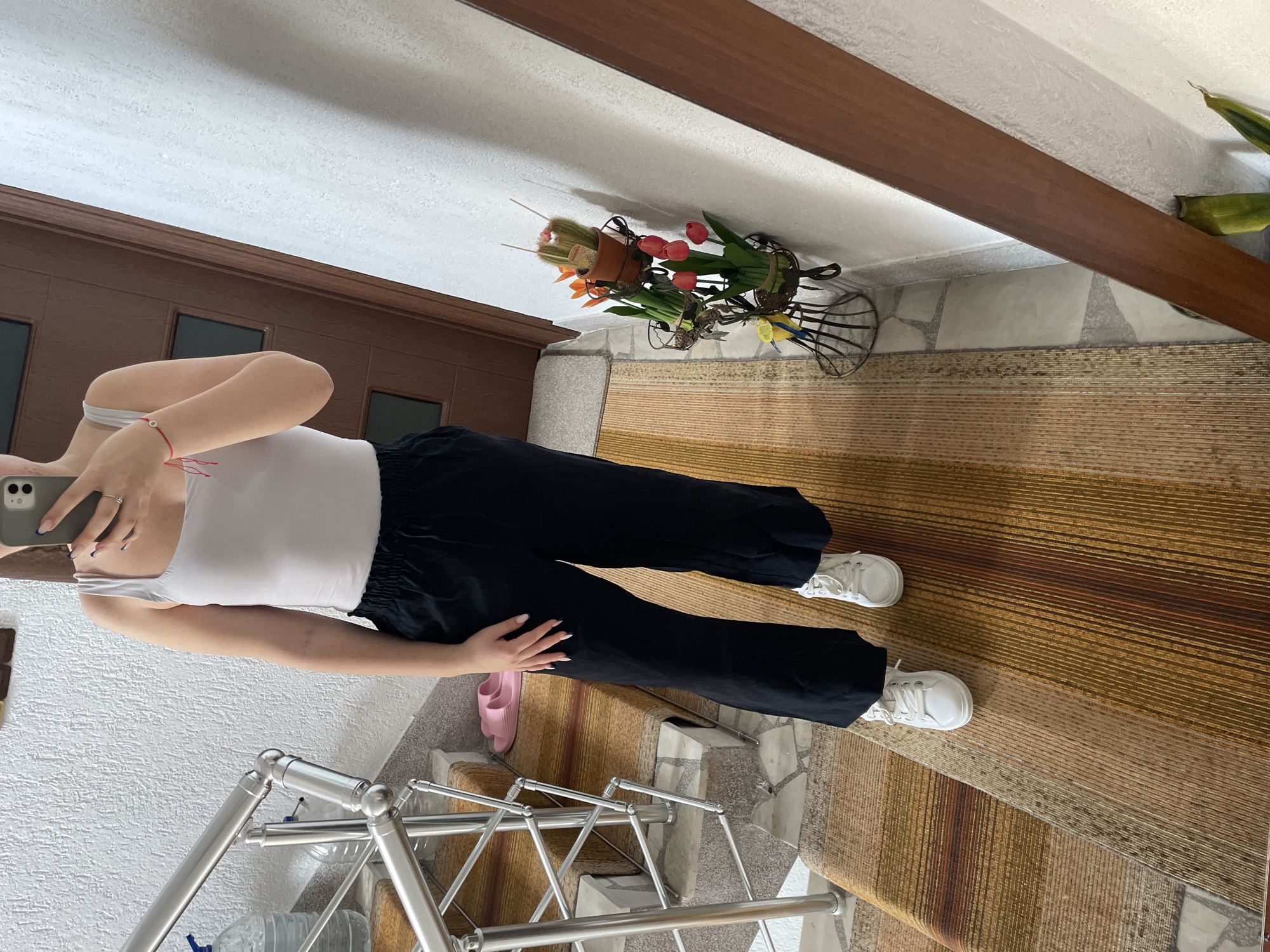 A person wearing a white tank top and black pants stands on a staircase. The staircase is covered with a patterned carpet, and there is a metal railing. Pink slides are visible near the person's foot. The background includes a door, a mirror with a brown frame, and a floral arrangement. The location is unknown.