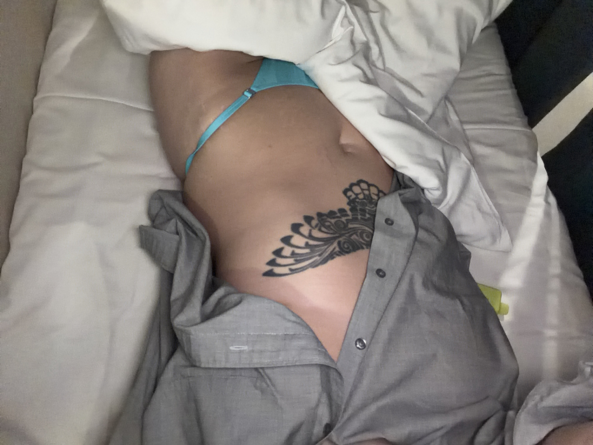 A person with a tattoo is lying in bed with an unbuttoned gray shirt and blue underwear. The location is unknown and cannot be determined from the image.