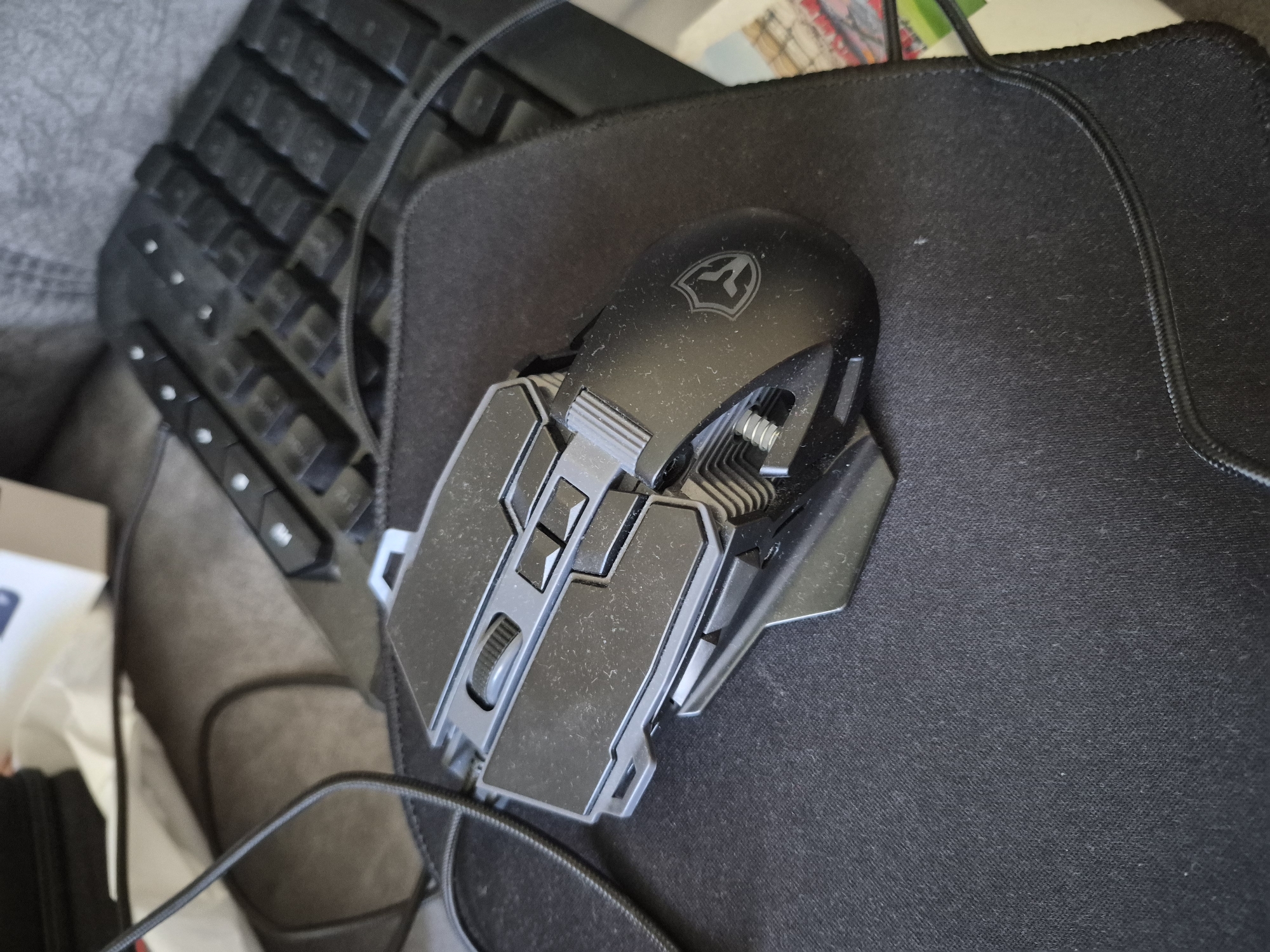 This is a close-up, high-angle, eye-level shot of a black gaming mouse on a black mousepad. The mouse is the primary subject, positioned diagonally from the bottom left to the top right of the frame. It features a textured surface, a scroll wheel, and several buttons. A stylized logo, resembling a shield with an angular design, is visible on the top of the mouse. The mousepad has a woven texture and a stitched edge.
Behind the mousepad, a portion of a black keyboard is visible, with its keys showing a grid-like pattern. The background also includes a textured gray surface, possibly fabric, and a hint of a colorful image or book in the upper right corner. A black braided cable snakes from the mouse, across the mousepad, and out of view. The lighting is bright, suggesting an indoor setting during the day, but there are no definitive external cues for time of day or weather. The scene appears to be a desk setup for gaming or computer use, with no people or direct action depicted. The overall impression is one of a still-life arrangement of computer peripherals.