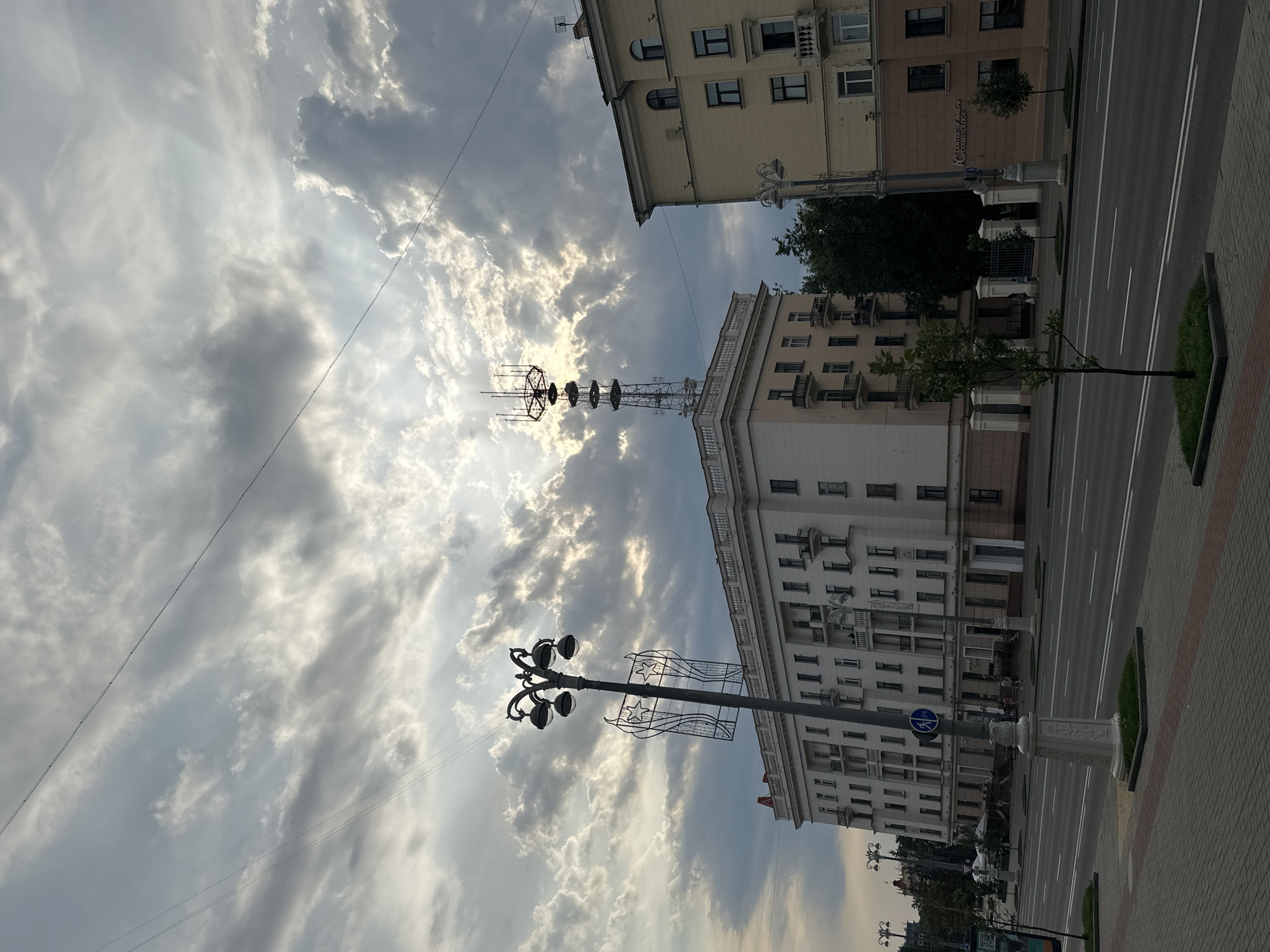 The image captures a view of the city of Minsk, Belarus, featuring a wide, multi-lane road in the foreground. On the left side of the road, a sidewalk is paved with gray and red bricks, bordered by small patches of grass. A decorative lamppost with multiple globes stands prominently on the sidewalk. Behind the sidewalk, a large, multi-story white building with numerous windows and balconies stretches across the frame. To the right of this building, a tall, lattice-like telecommunications tower rises above the roof. Further to the right, a yellow building with arched windows and balconies is visible, with a street lamp in front of it. The sky above is dramatic, filled with a mix of dark and light gray clouds, with rays of sunlight breaking through. A few trees are scattered along the buildings. The scene appears to be a public street in an urban environment.