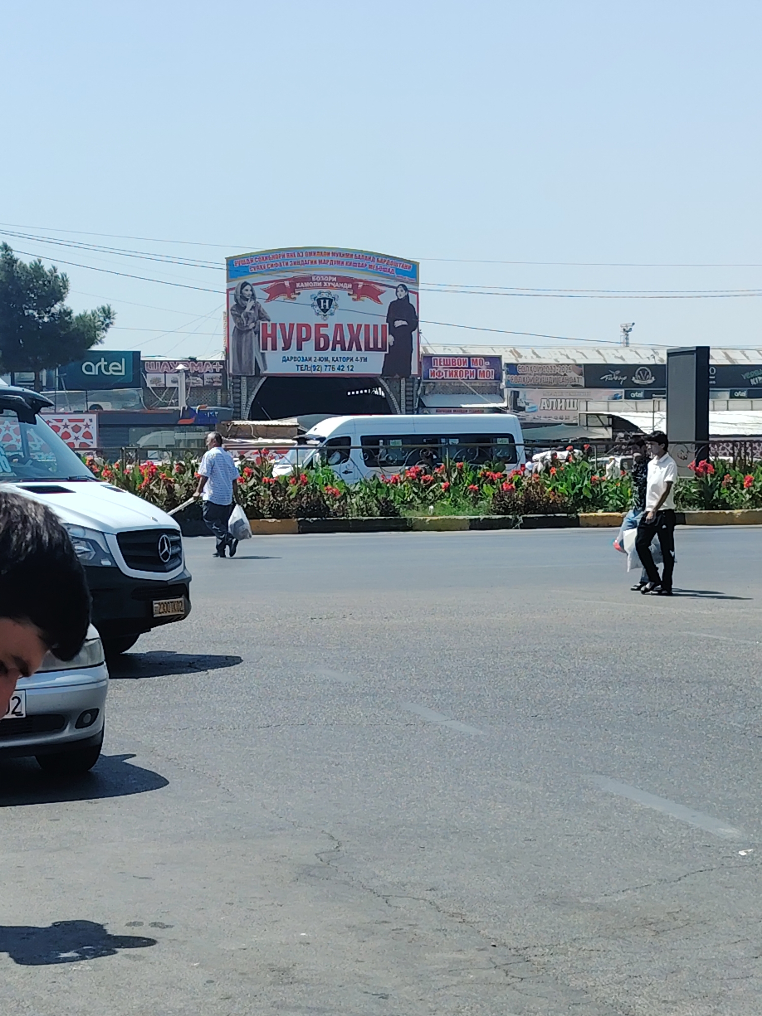 The image captures a daytime street scene in Ghafurov, Tajikistan, with a large billboard prominently displaying text and an image of a woman in traditional attire. The billboard's text is in Cyrillic and appears to advertise a business named "НУРБАХШ" with a phone number listed as "(92) 776 42 12". In the foreground, a white Mercedes-Benz van is parked, with part of a silver car visible on the left. A man in a checkered shirt and dark pants walks away from the van, carrying a white plastic bag. Further down the street, a white minibus and another van are visible. Two individuals, one in a white shirt and black pants and the other in a patterned shirt and dark pants, are walking across the street, also carrying bags. Red flowers and green foliage line the median of the road. The overall environment suggests a commercial or bustling area within the city.