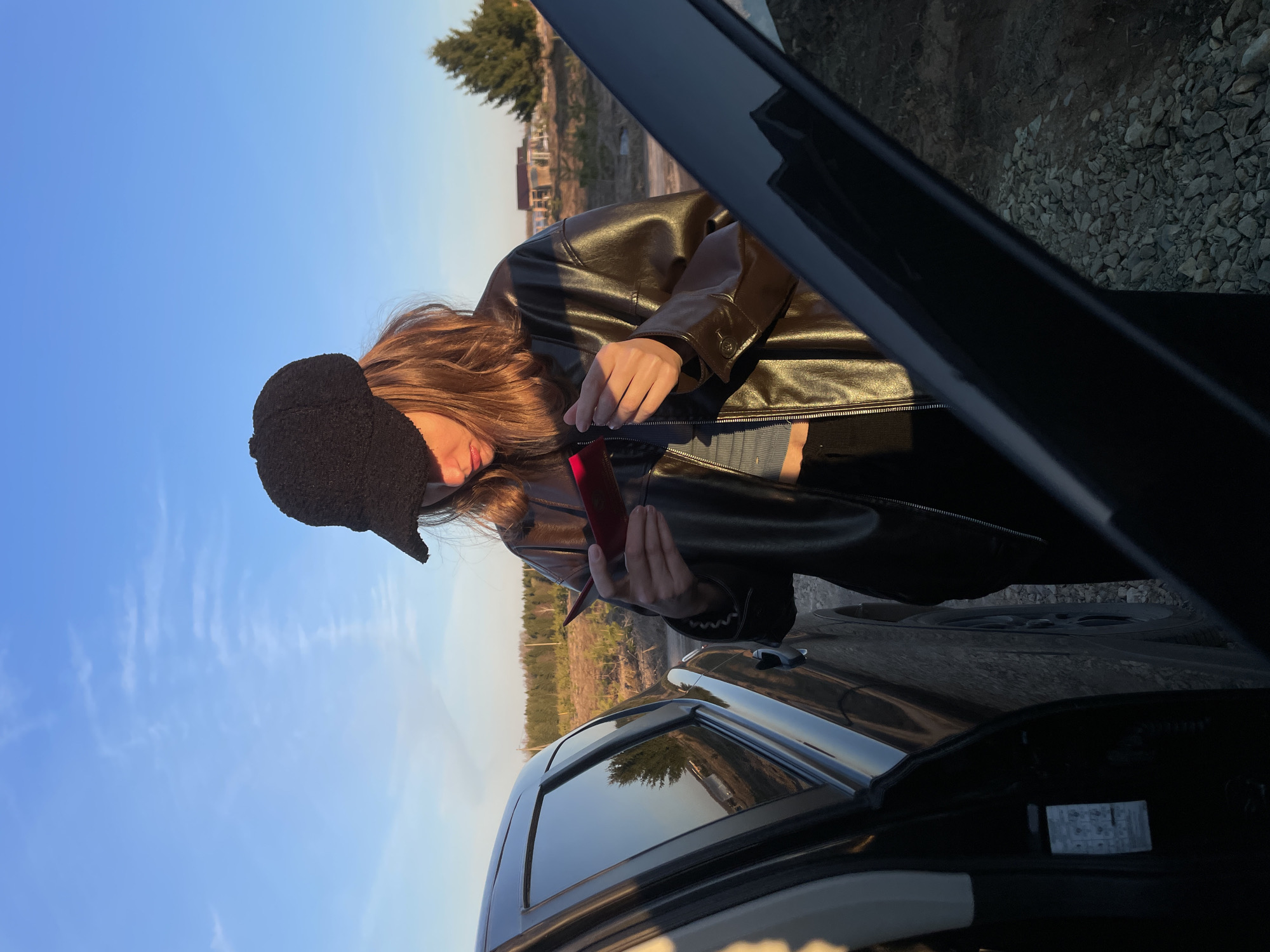 The image depicts a young person standing next to an open car door in an outdoor, semi-rural setting, consistent with Подшивалово, Russia. The person has long, brown hair and is wearing a glossy brown leather jacket over a lighter top, and a dark, textured hat. Their head is tilted down, and they are focused on a bright red, rectangular object held in their hands.
The scene is bathed in the warm, golden light of late afternoon or early evening, casting soft shadows. The sky above is a clear, vibrant blue with a few faint, wispy clouds. The person is positioned beside a dark-colored car, which has its driver's side door open, revealing a glimpse of the interior. The car's side window reflects the bright sky and distant landscape.
The background shows a horizon line with an expanse of trees and sparse vegetation, indicating a natural environment. Beyond the immediate foreground, faint outlines of distant buildings or structures are visible. The ground directly beneath the car and person appears to be a gravel or dirt surface. No visible text is legible in the image. The overall impression is a quiet moment captured outdoors, perhaps during a stop on a journey.